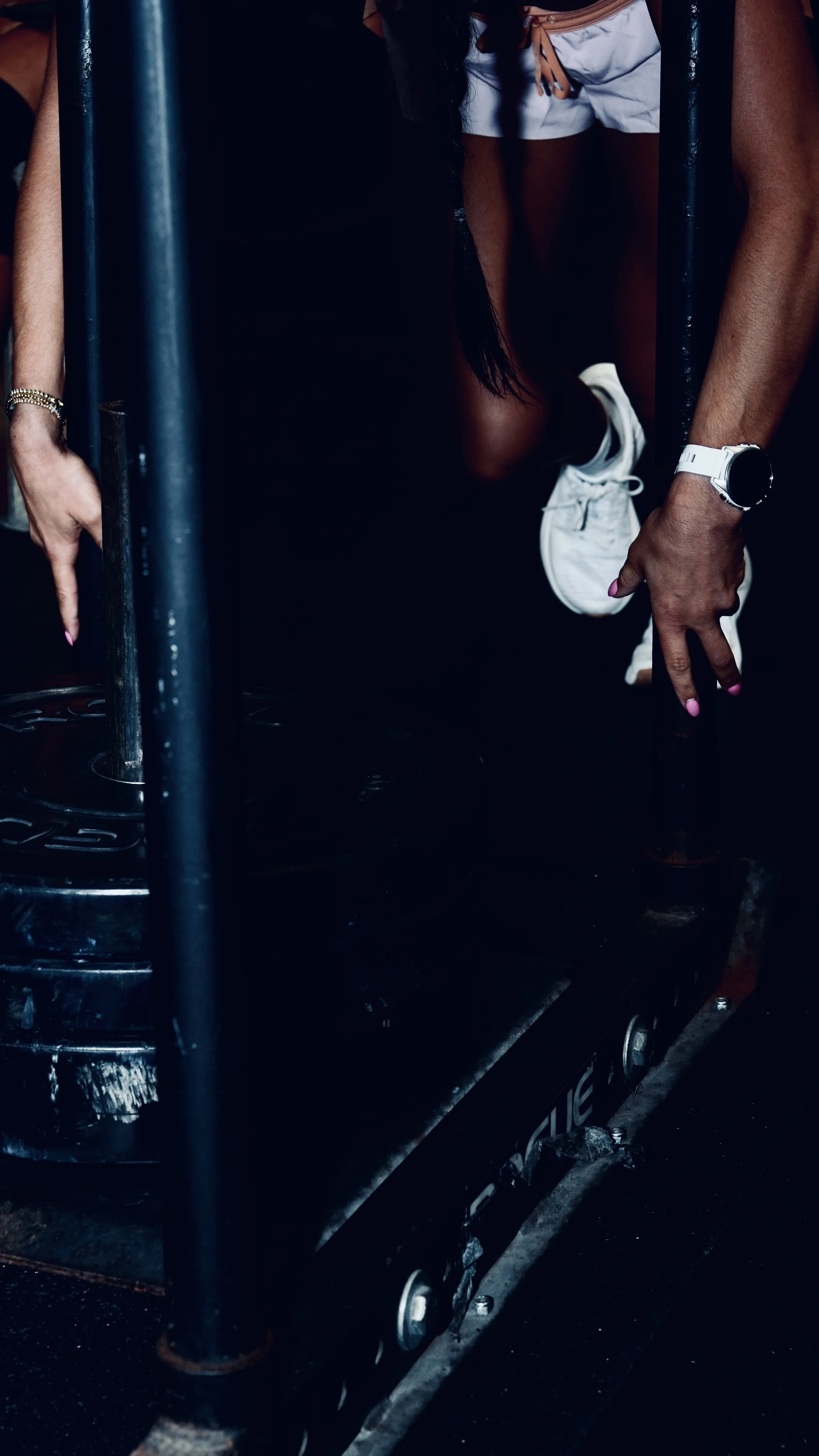 Close-up of a person wearing a white watch and shorts, with their hand resting on a black gym equipment, in a dimly lit setting.