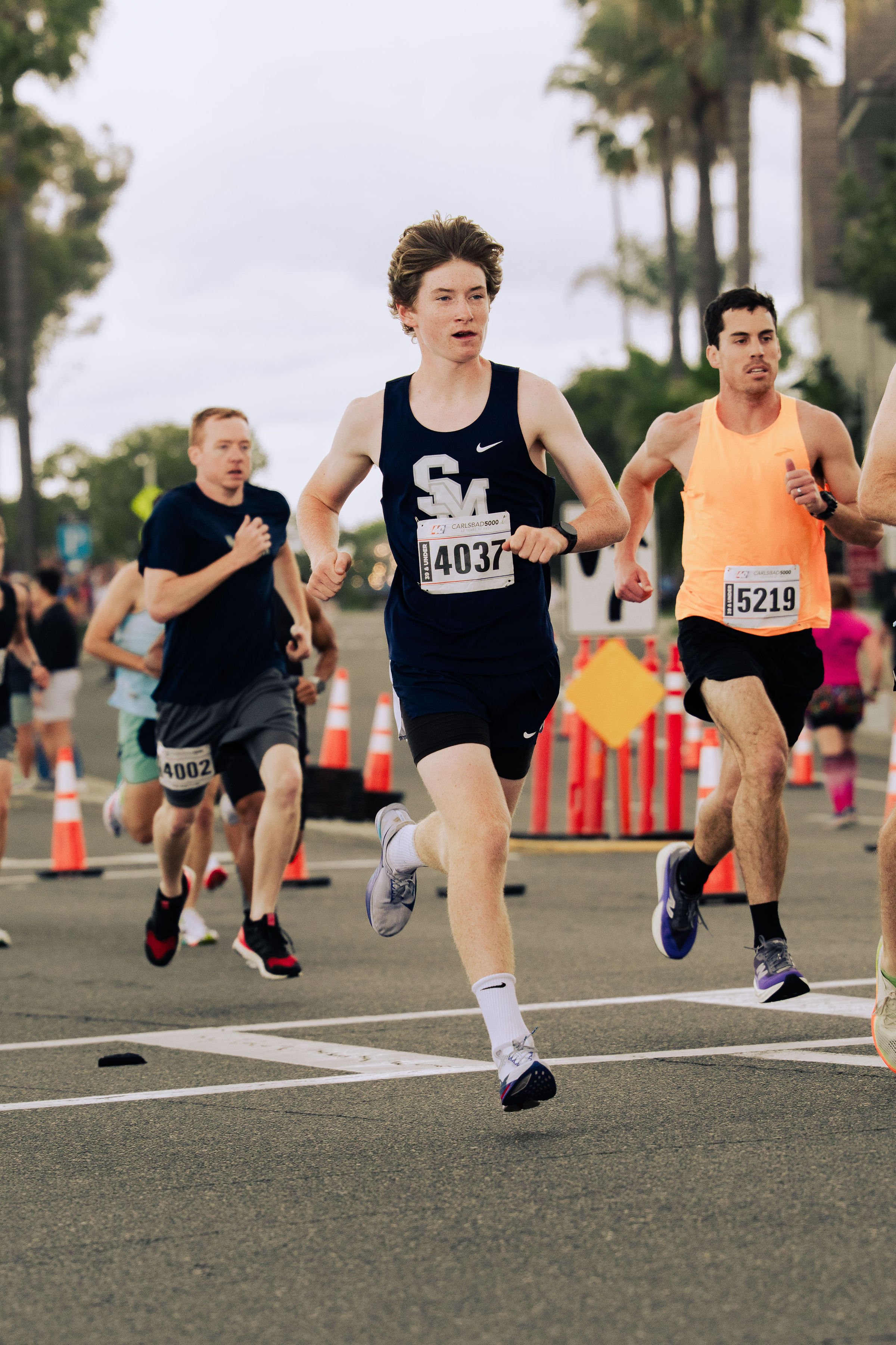 Multiple runners competing in a road race, with some wearing bib numbers and athletic gear, on a city street lined with orange cones and trees, under an overcast sky.