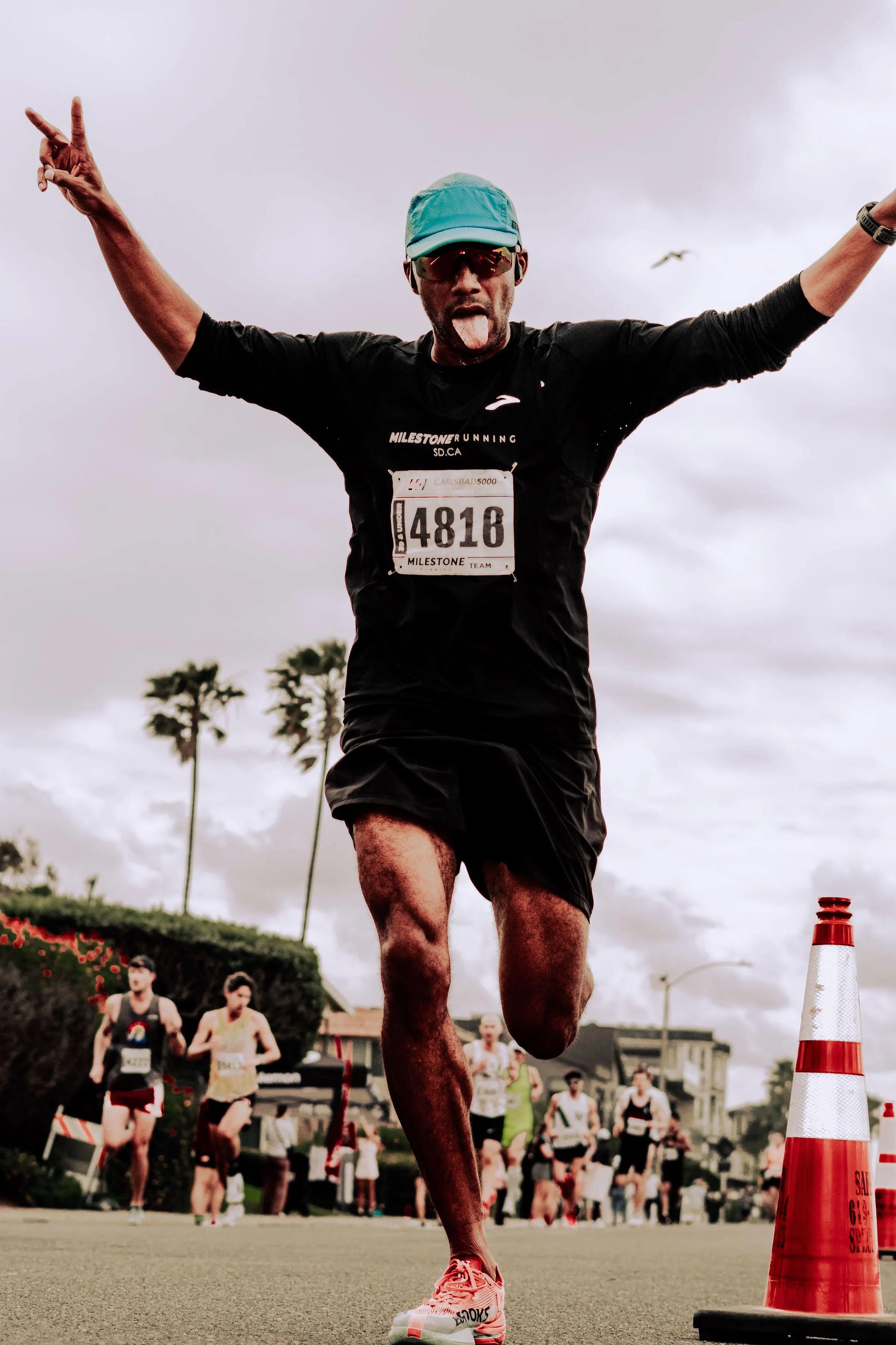 A male runner wearing a teal cap, sunglasses, and black athletic clothing crossing the finish line with a race bib number 4816. He is sticking his tongue out and raising his arms in celebration. Several other runners are visible behind him, along wit