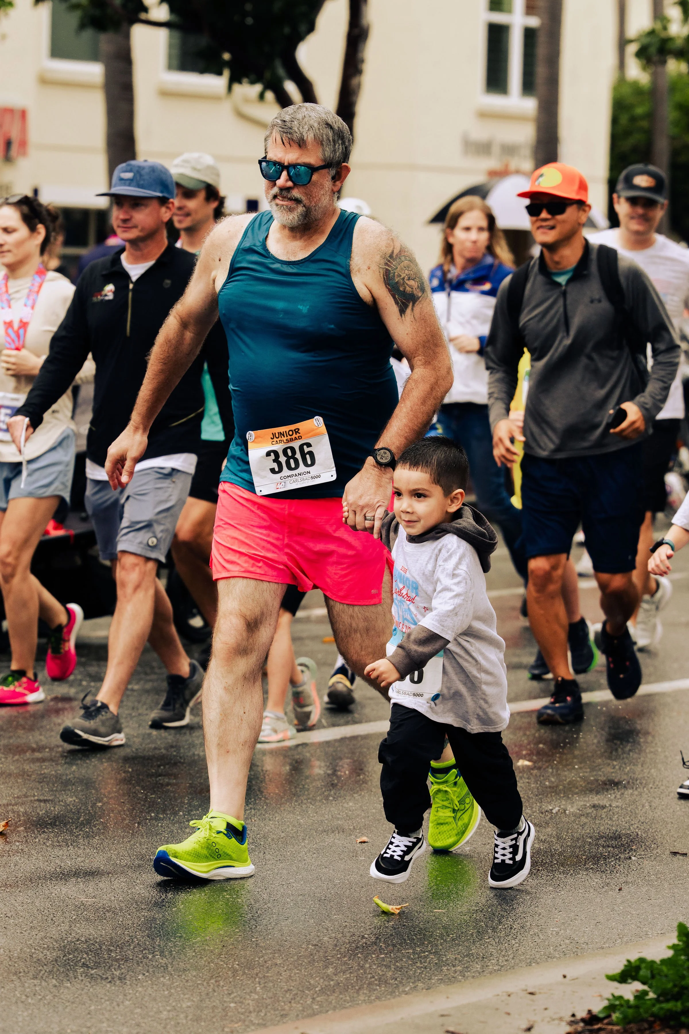A man and a young boy holding hands while participating in a running event on a wet street, with other runners and spectators in the background.