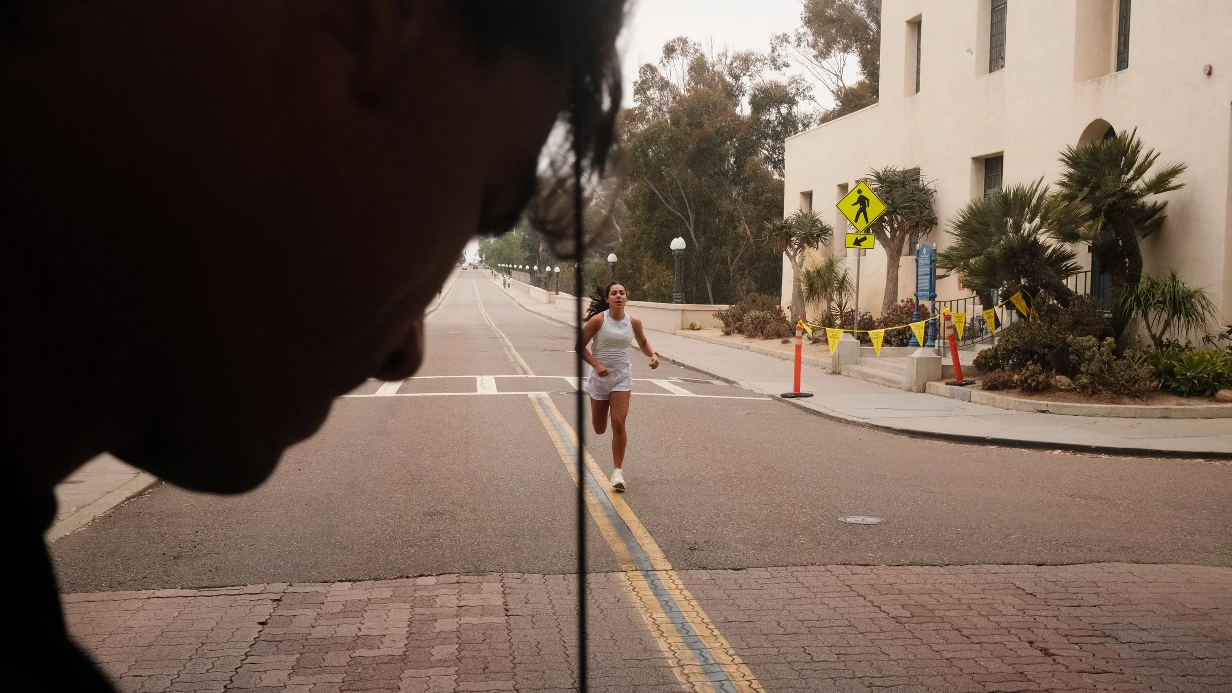 A woman running on a city street as seen through a window or glass, with a person's face partially visible in the foreground.