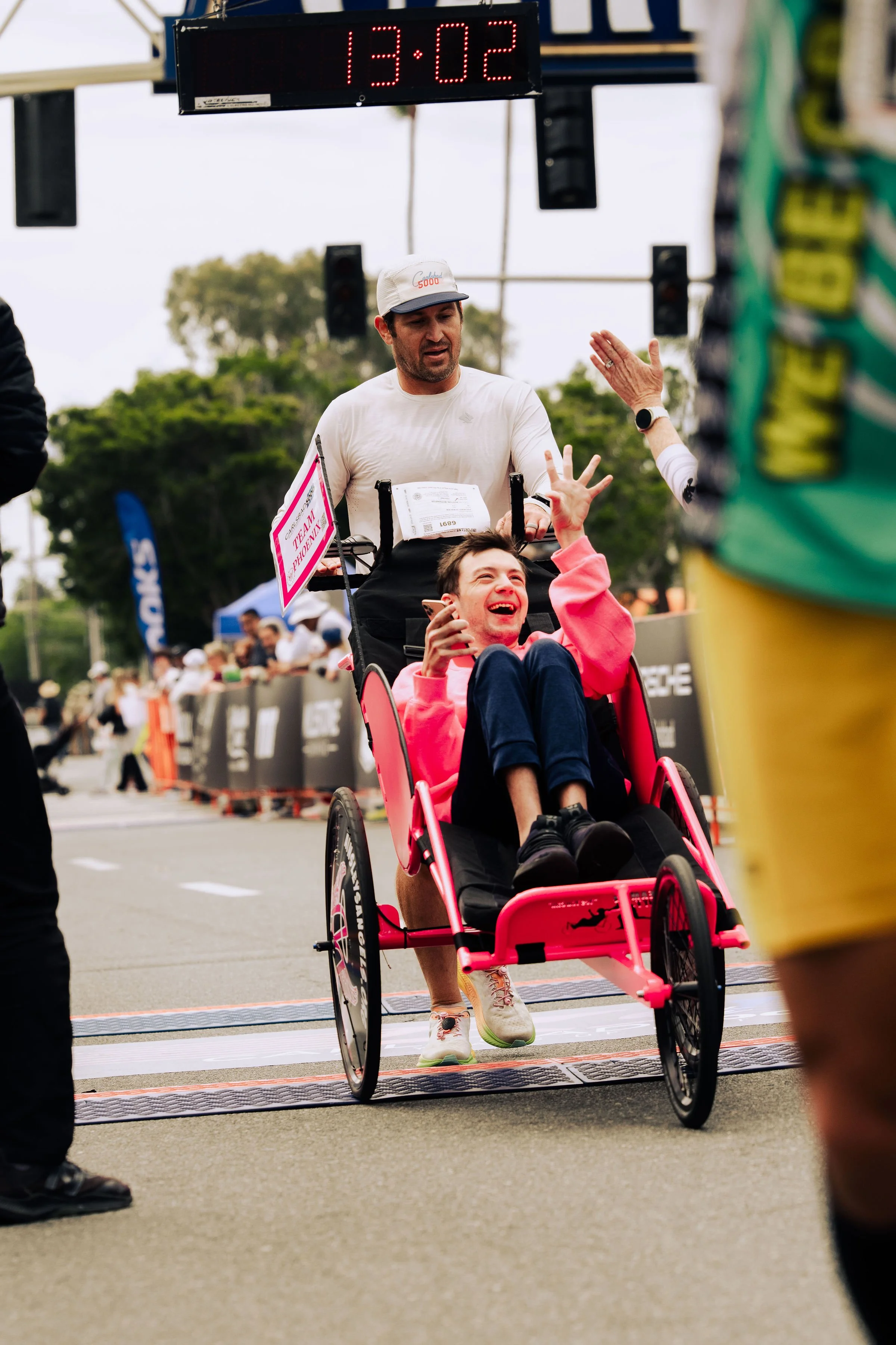 A man in a white shirt and white cap pushing a woman in a pink wheelchair across a finish line, with a digital timer showing 13:02 overhead and a crowd in the background.