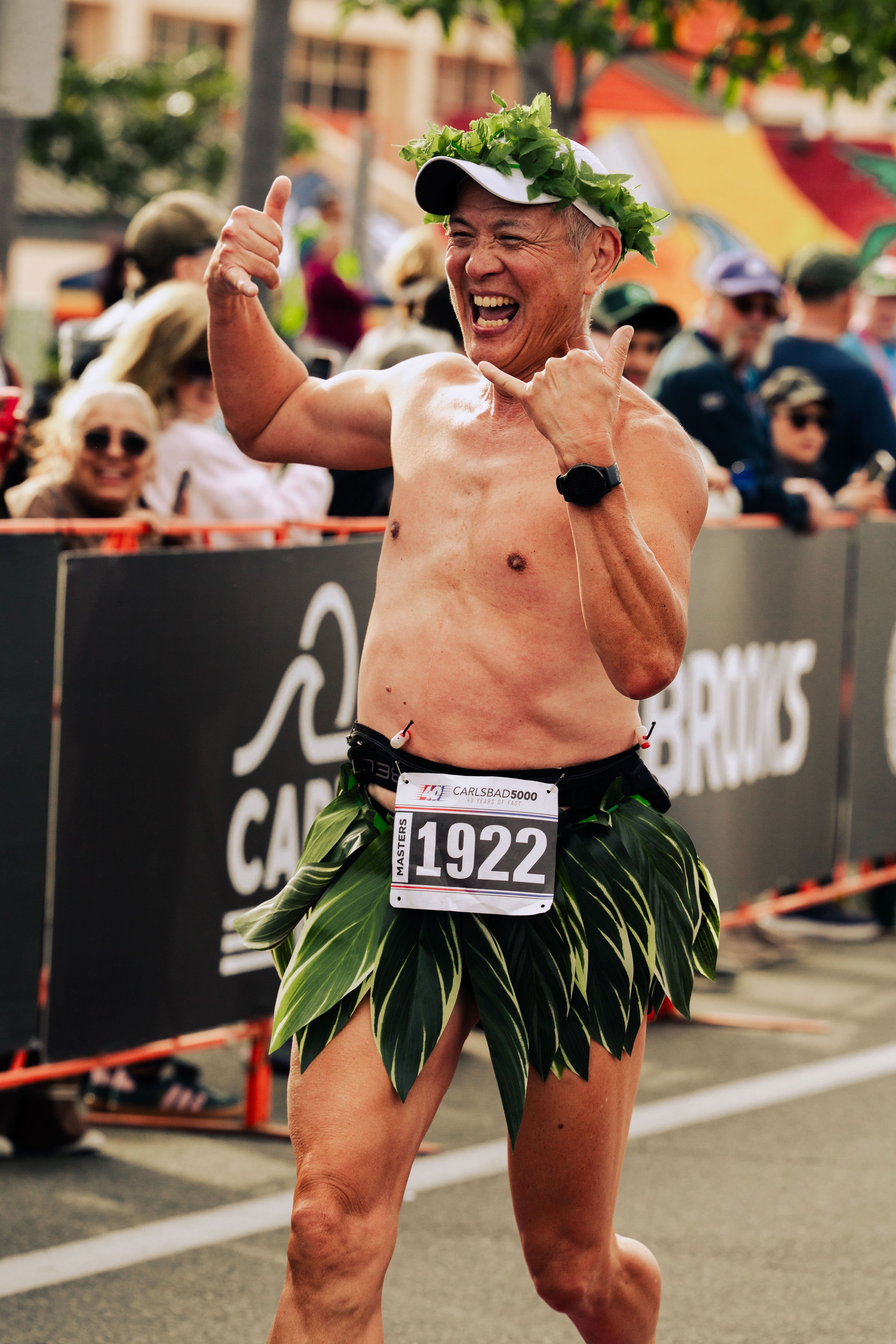 An elderly man smiling and celebrating at a marathon event, dressed in a grass skirt with a race bib number 1922 and wearing a laurel crown and a baseball cap.