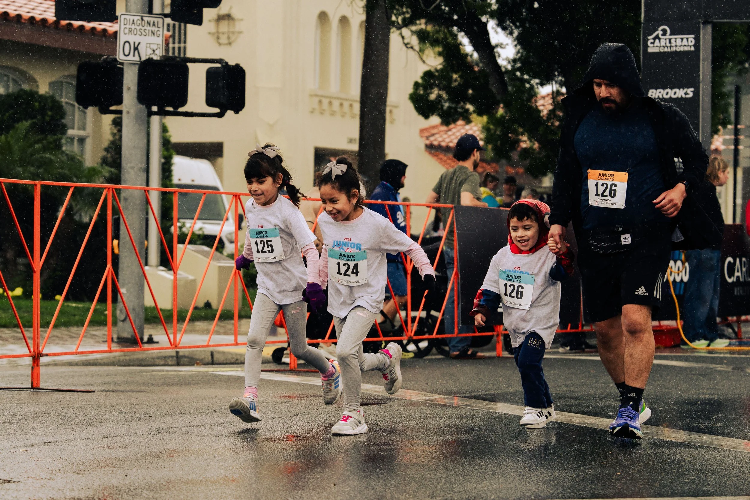 Children running in a race during rainy weather, with race bibs on their shirts and a man holding one child's hand. There are orange barricades, a street sign, and a Carlsbad California sign in the background.