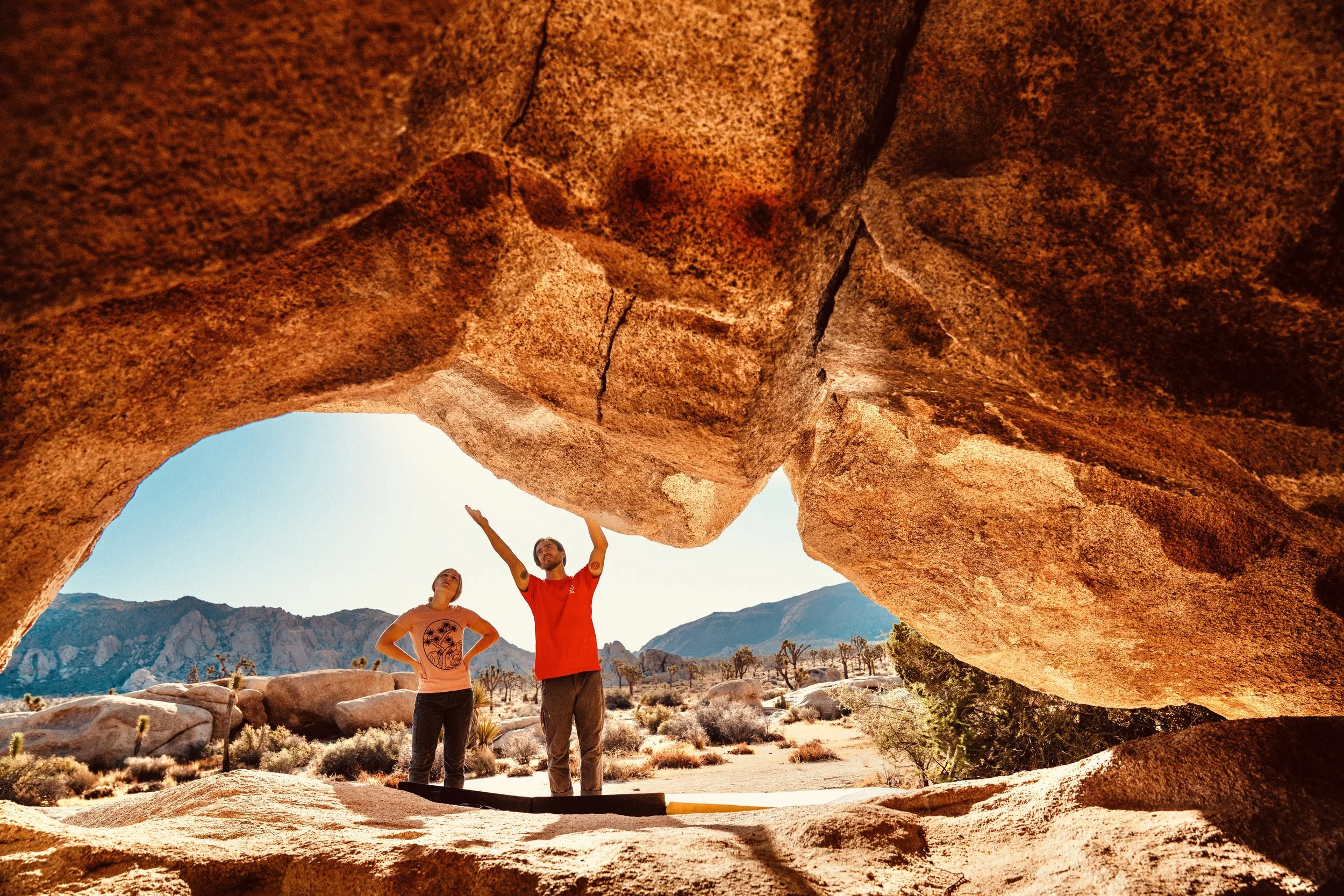 Two people standing under a large, natural rock formation in a desert landscape with mountains in the background, during daytime.