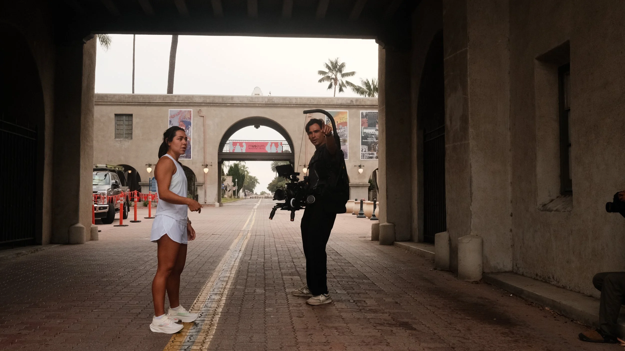 A woman in a white outfit and sneakers talking to a man with a camera on a gimbal, under an archway in an urban area with palm trees and posters on the wall in the background.