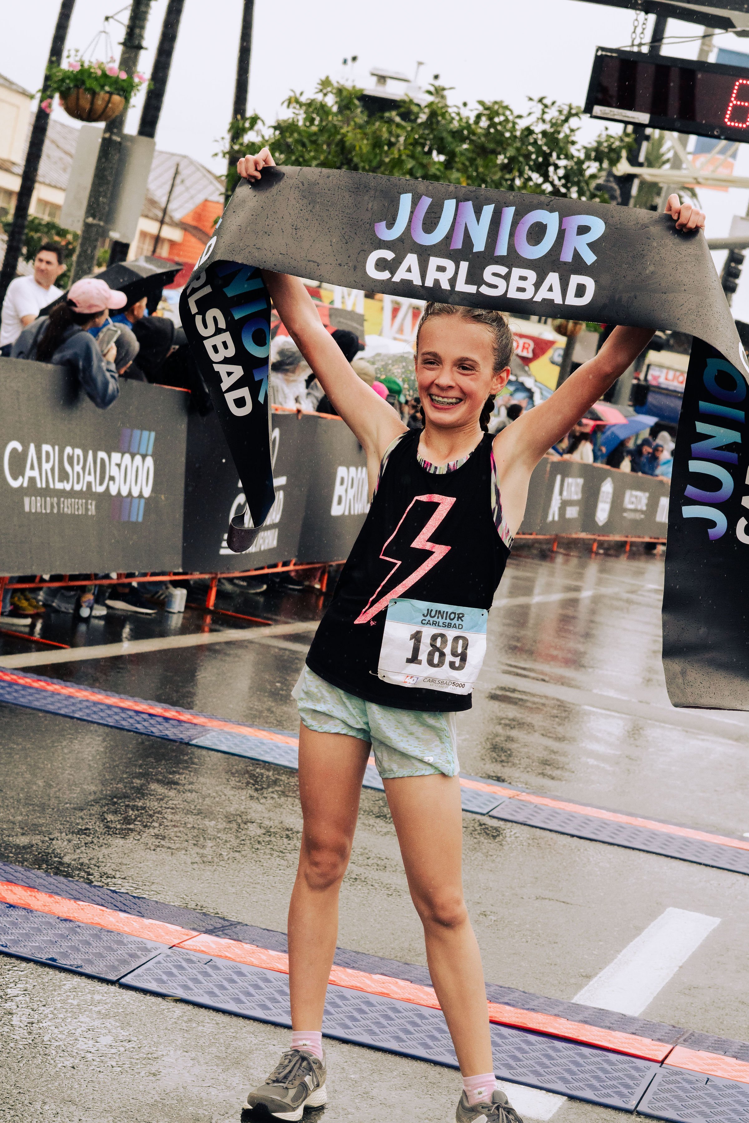 Young girl smiling and raising a race medal above her head after finishing a running race at Carlsbad race event on a rainy day.