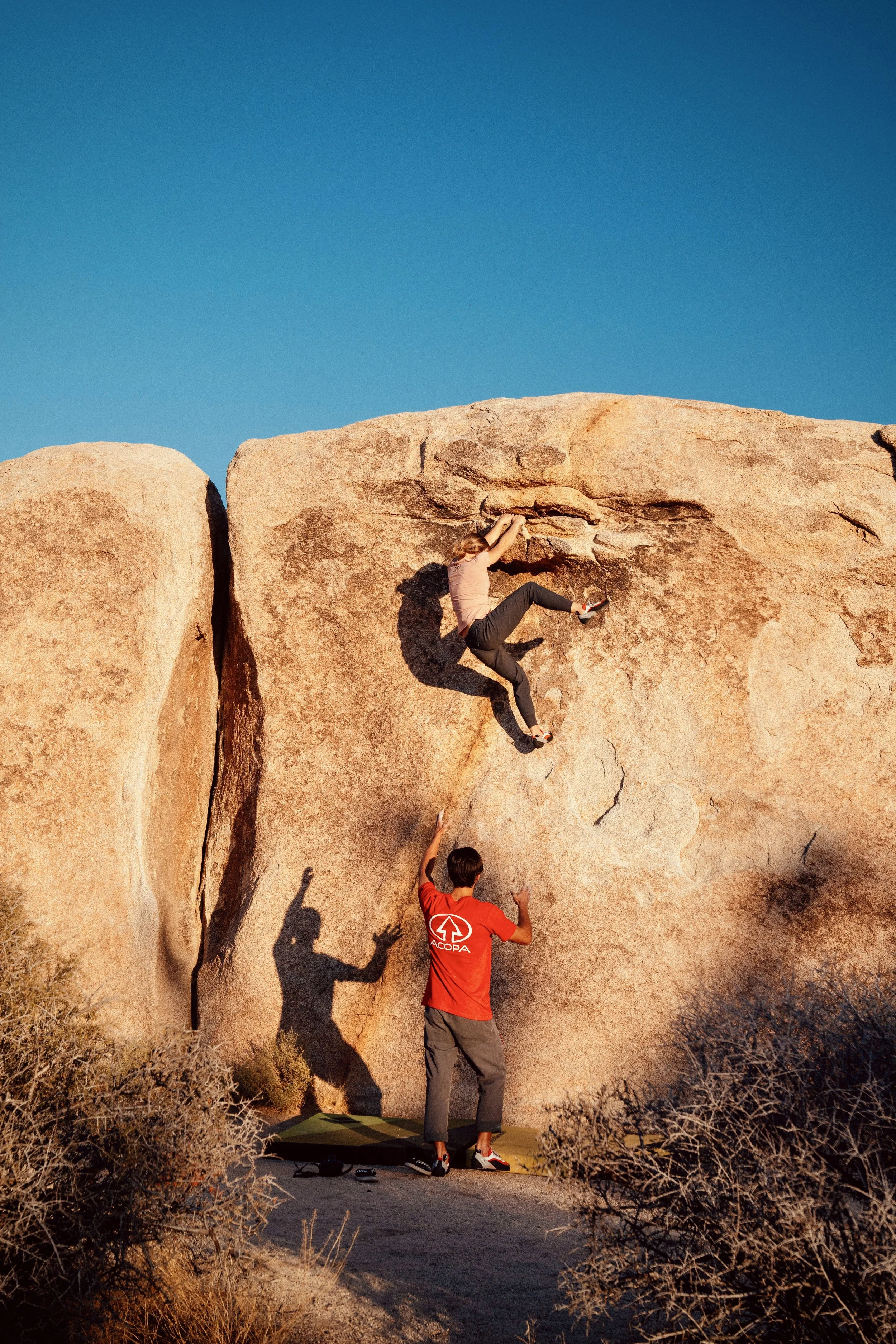 A woman rock climbing on a large, rugged rock formation under a clear blue sky, assisted by a man standing at the base, with dry desert shrubs in the foreground.