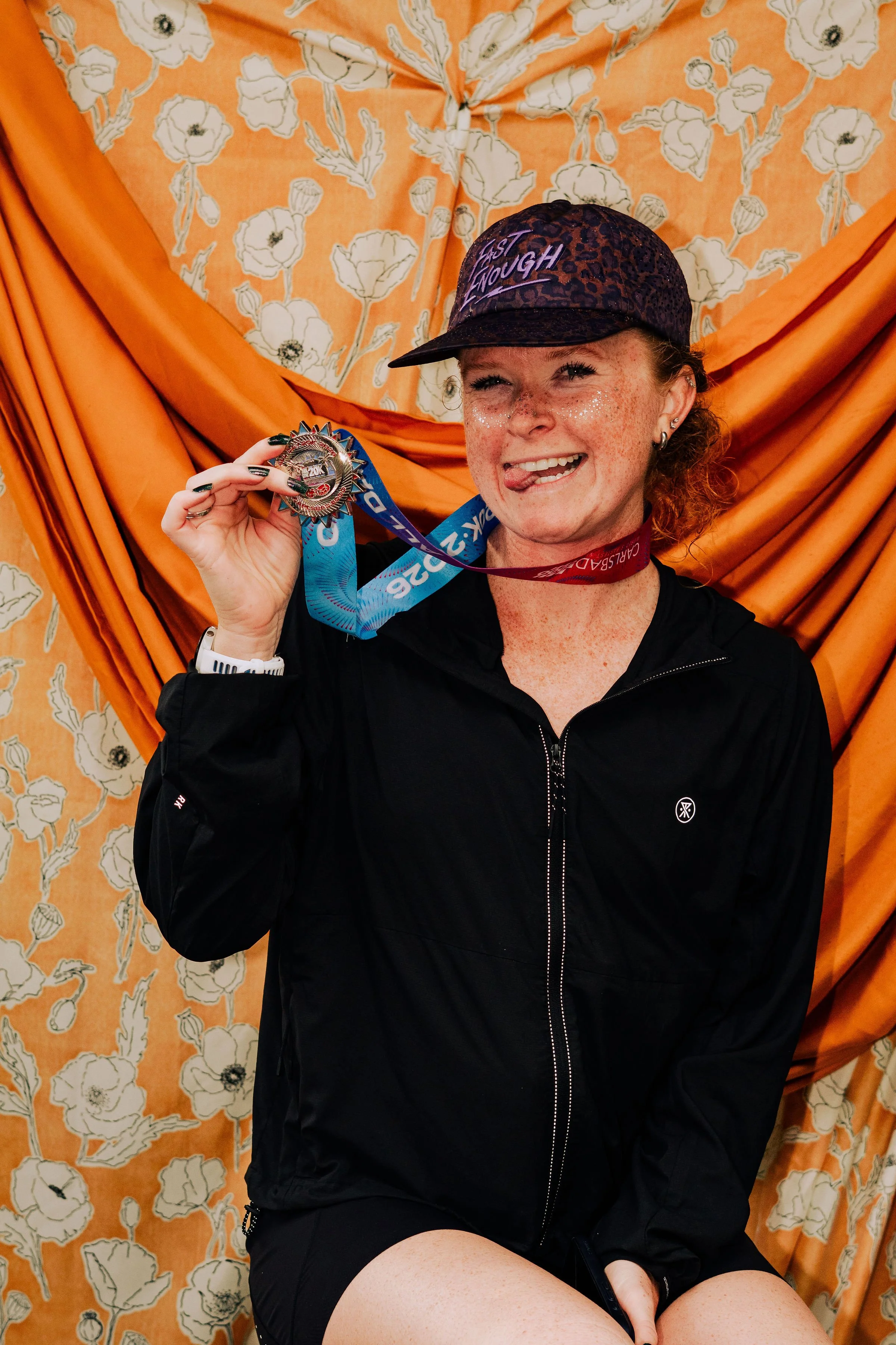 A woman with freckles and curly hair, wearing a leopard print cap with the words 'Last Enough,' is smiling and holding a medal. She is dressed in a black jacket and shorts, sitting in front of an orange floral fabric backdrop.