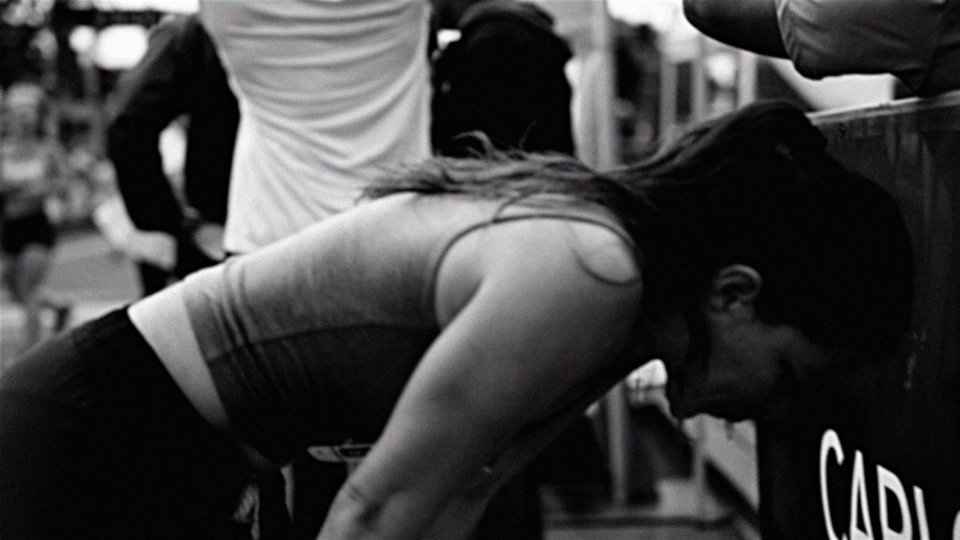 A woman is bent over, leaning on a table or counter. She is wearing a sleeveless top. There are other people around, and the background is blurred.
