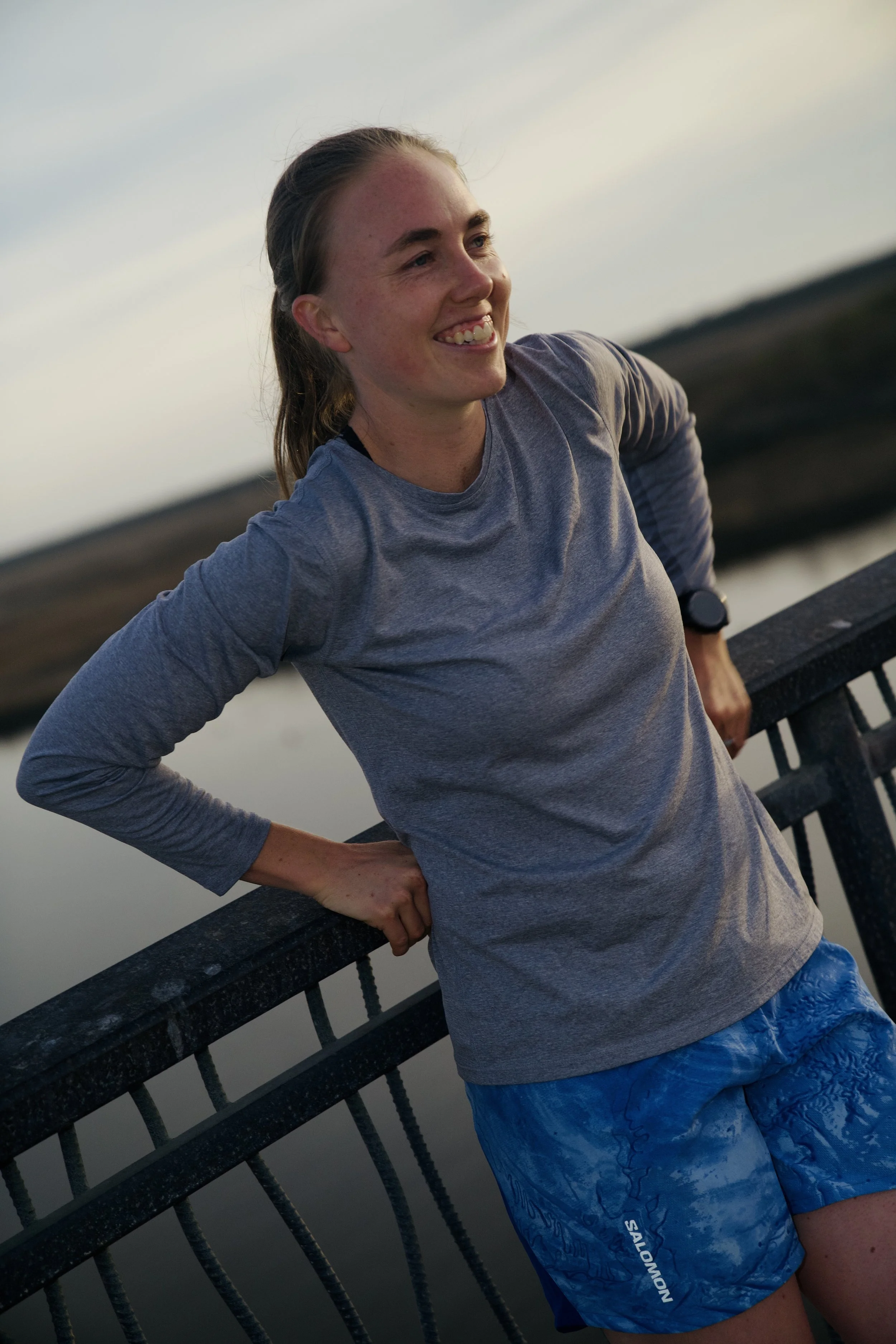 A young woman with long hair smiling, standing outdoors on a bridge, wearing a gray long-sleeved shirt and blue shorts, with water and landscape in the background during the evening.