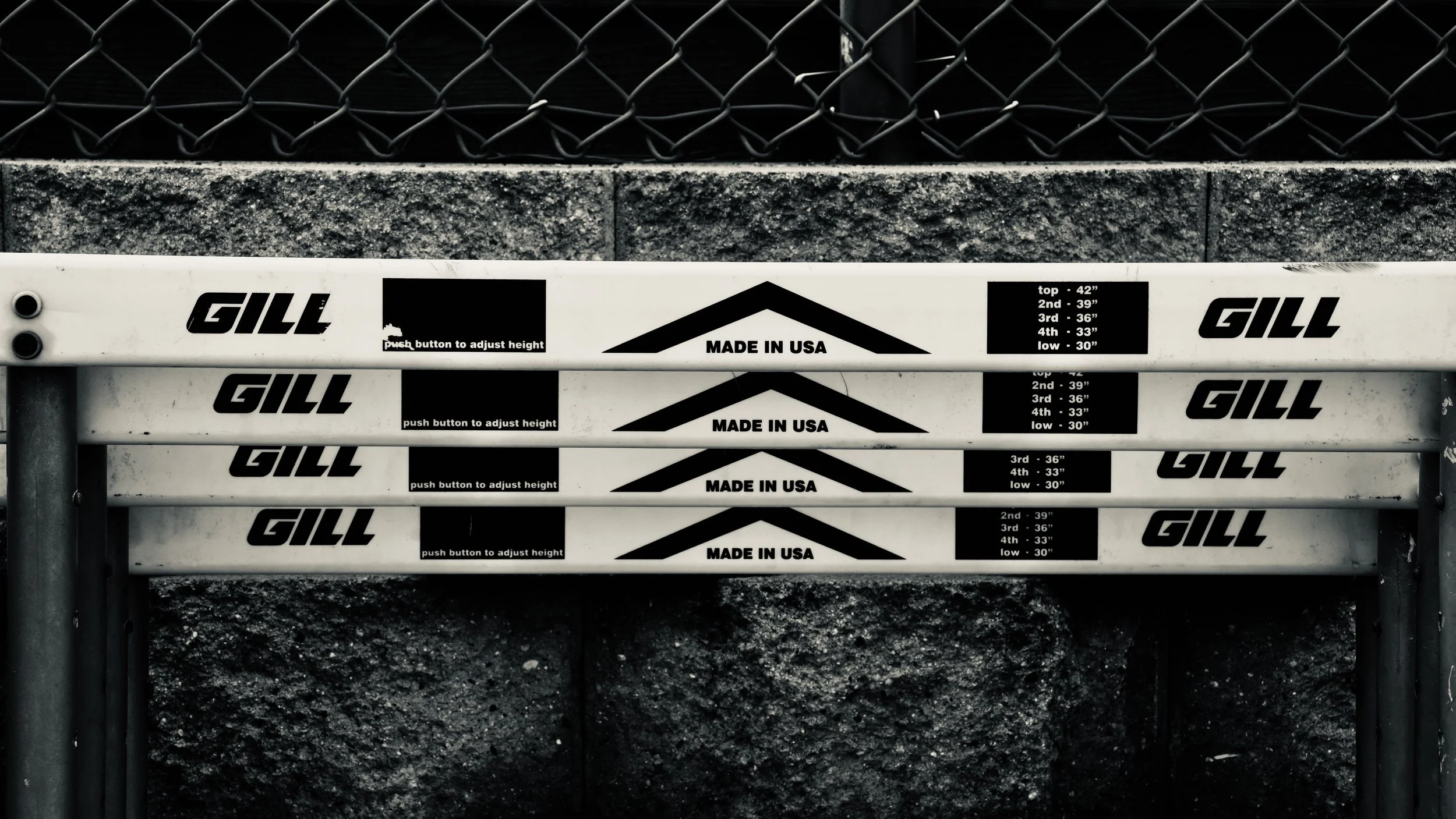 A stack of four white metal signs with black text and arrows, measuring different heights, placed against a gray textured wall and a black chain-link fence. The signs display brand names, size measurements, and instructions for height adjustment.