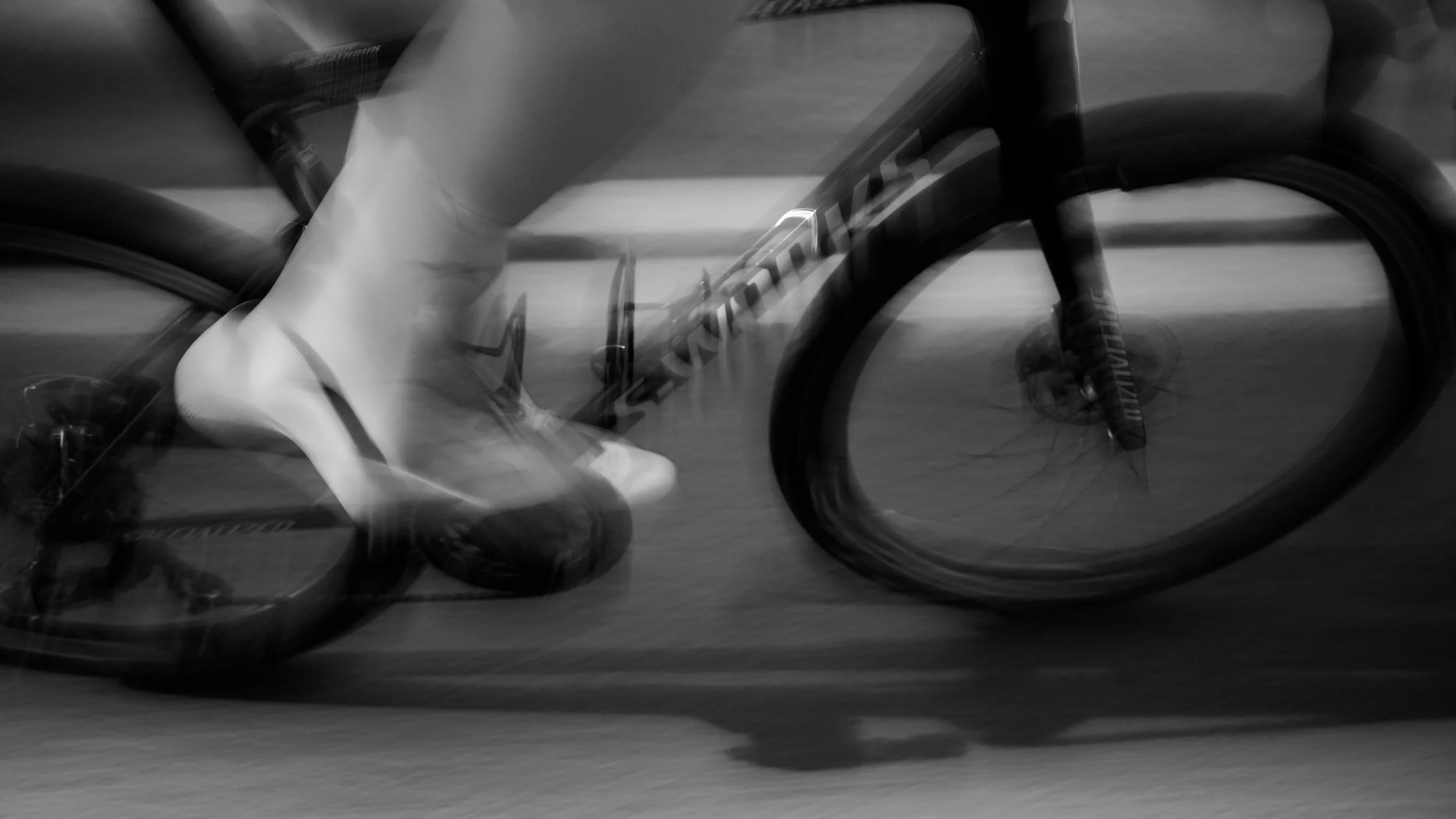 Black and white motion-blurred image of a person riding a mountain bike, with focus on the front wheel and part of the frame.