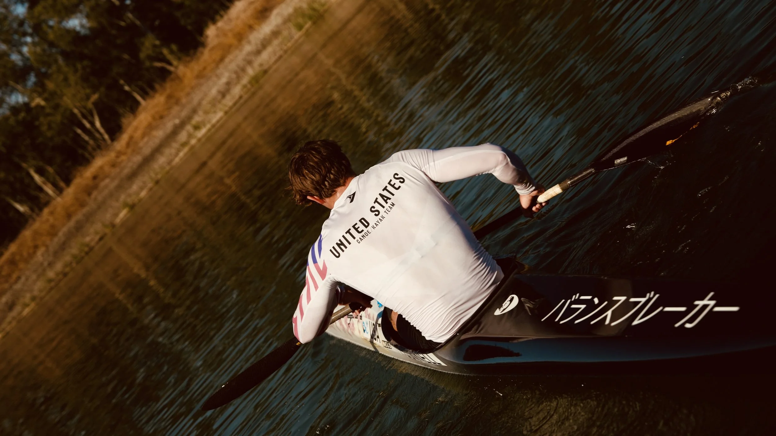 Person in a white long-sleeve shirt paddles a kayak on a body of water during sunset or sunrise, with trees and shoreline in the background.