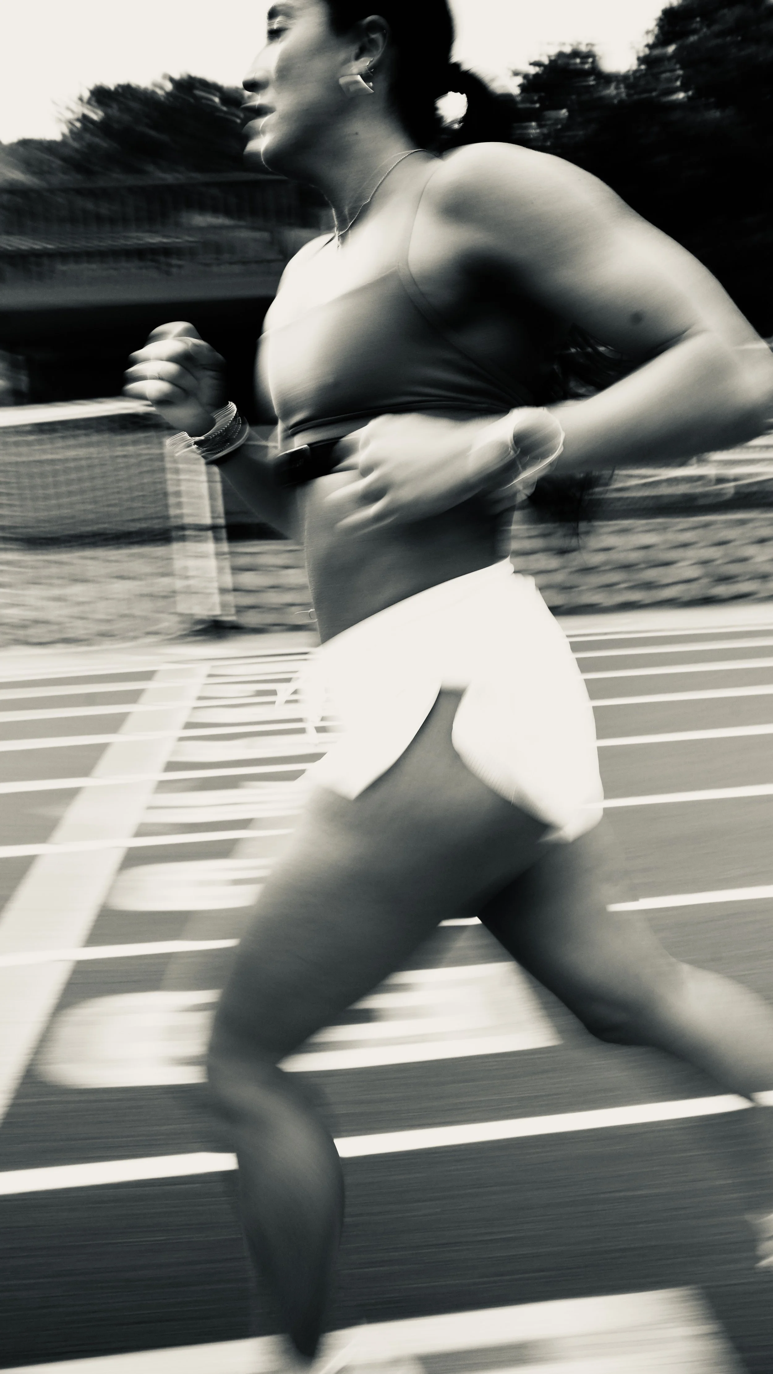 Blurred black-and-white photo of a woman running on a track, wearing a sports bra and shorts, with trees and a fence in the background.