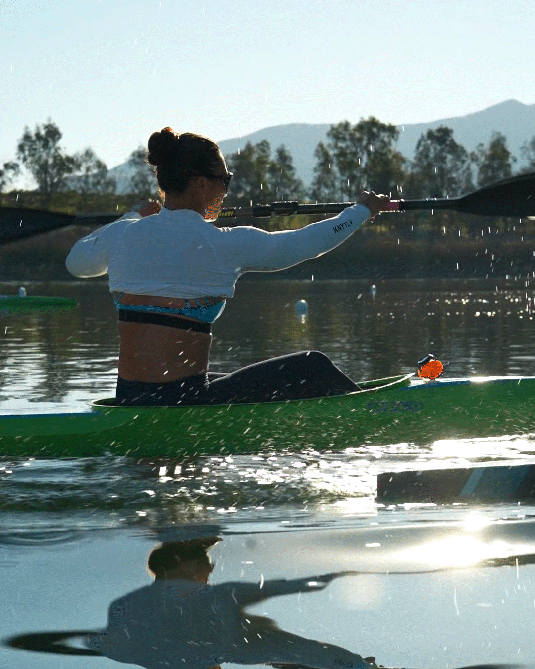 A woman kayaking on a sunny lake with mountains in the background, wearing a white long-sleeve top and sunglasses, holding a paddle, with her reflection visible in the water.