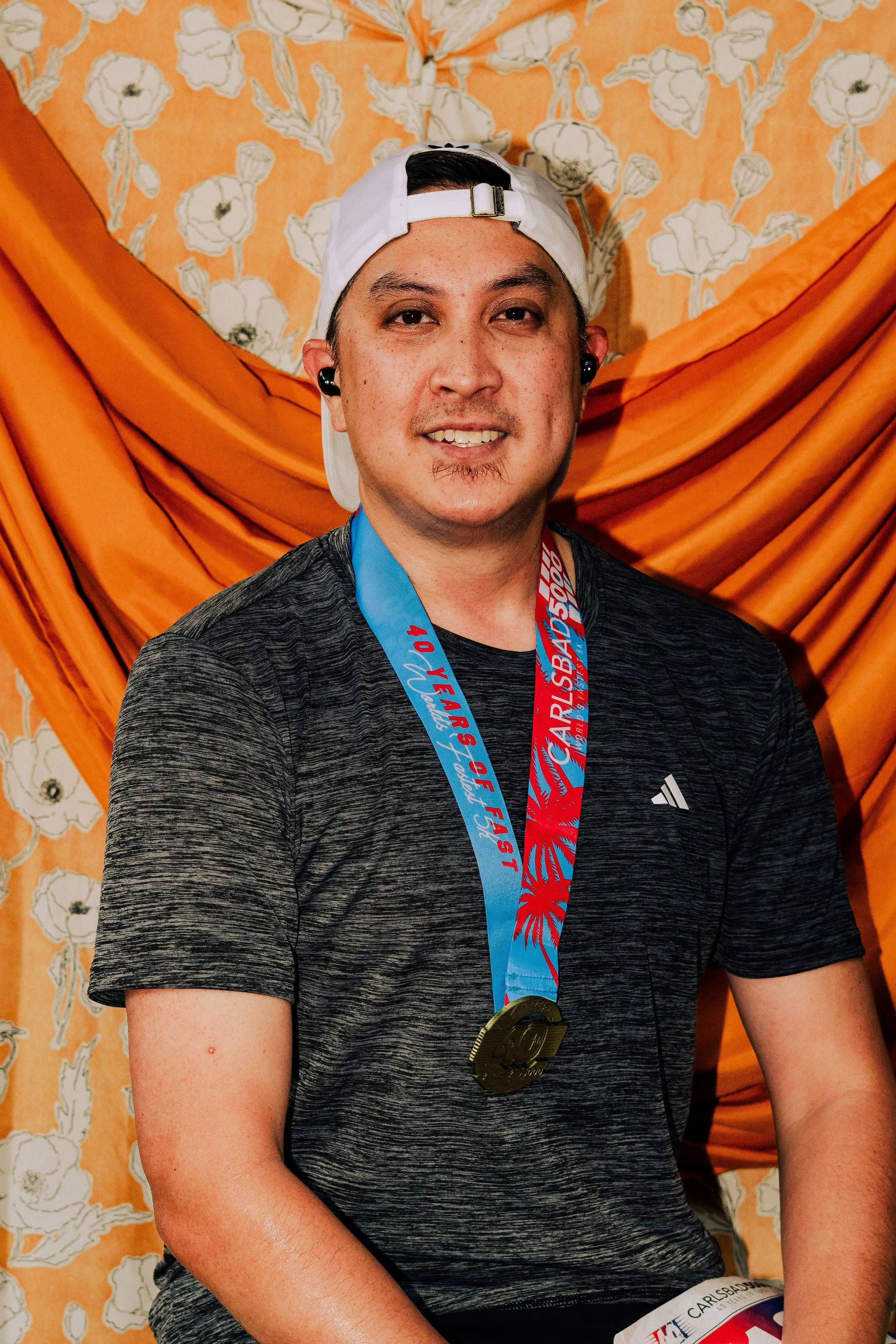 A smiling man wearing a white cap backwards, black earphones, and a dark gray athletic shirt. He has a medal around his neck and is seated in front of a decorative orange floral curtain background.