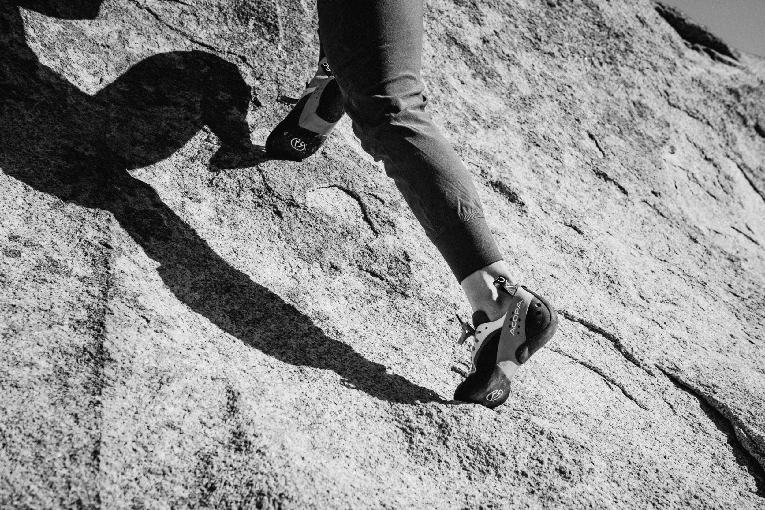 Close-up of a person's legs climbing a steep rock face, wearing gloves and climbing shoes, with a shadow cast on the textured rock surface.