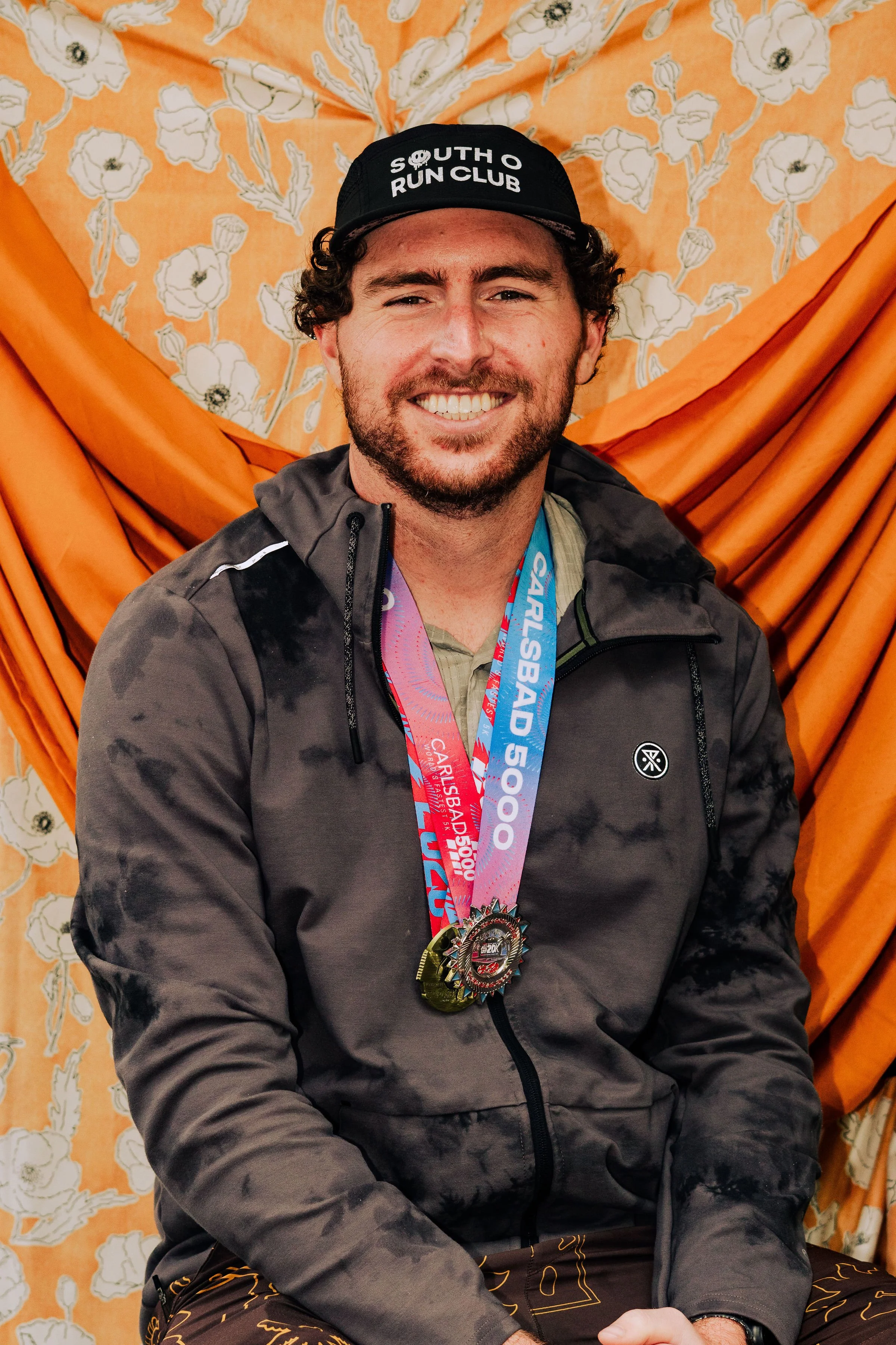 A man with dark curly hair and a beard smiling while wearing medals around his neck. He is dressed in a dark, patterned jacket and sitting in front of a floral orange backdrop with draped fabric.