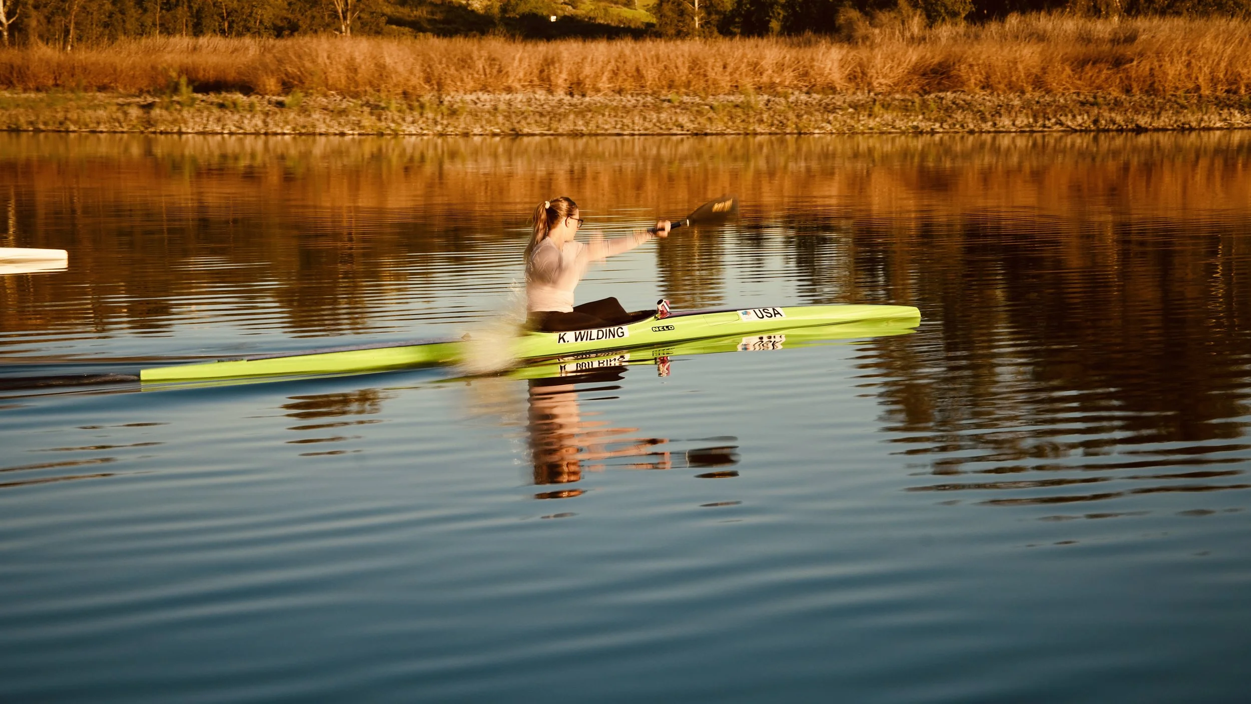 A woman in sunglasses paddling a lime green kayak on a calm body of water during sunset, with orange and brown foliage along the shoreline.