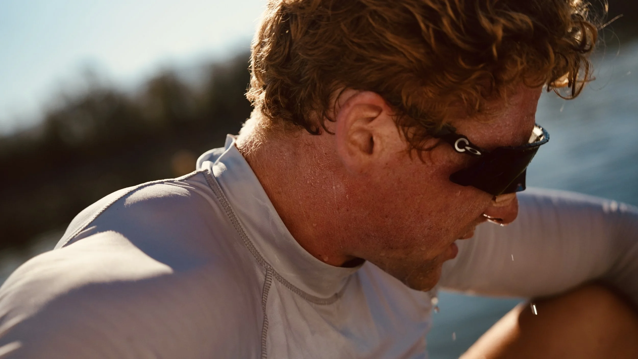 Close-up of a man with curly reddish hair wearing sunglasses and a white shirt, looking down, outdoors near water during the daytime.