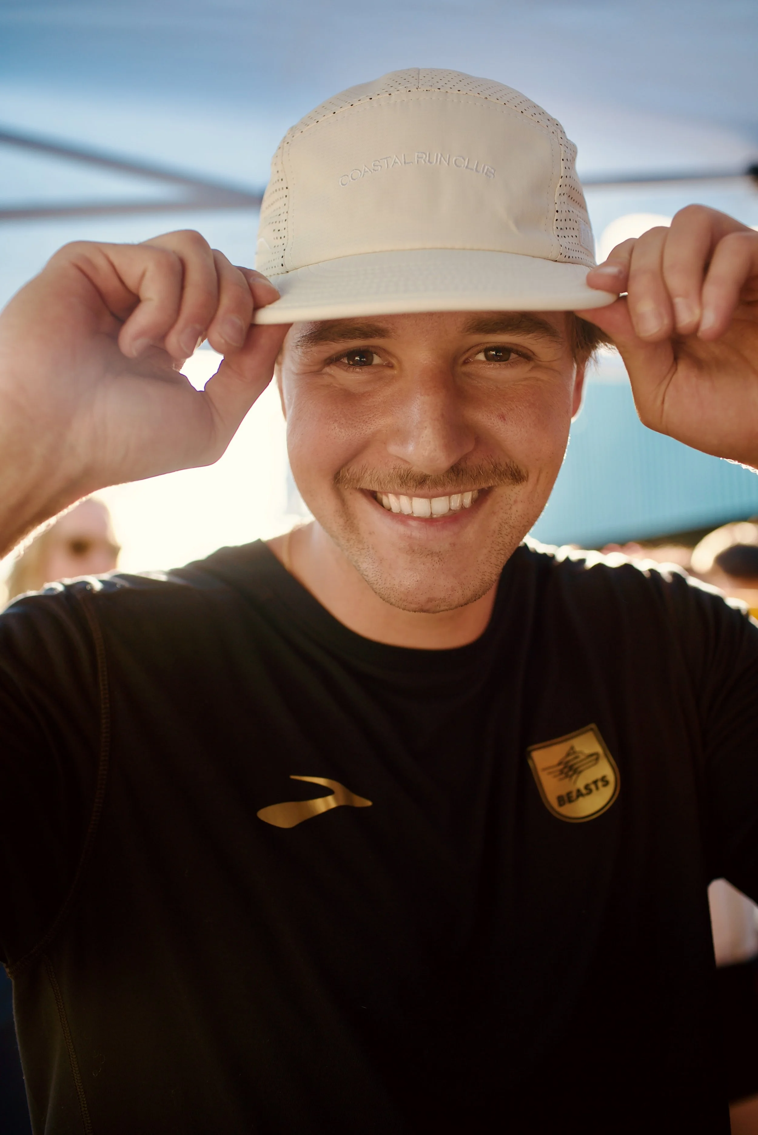 A man with light skin, brown hair, and a mustache smiling while lifting a light-colored cap with the words 'COASTAL RUN CLUB' embroidered on it.