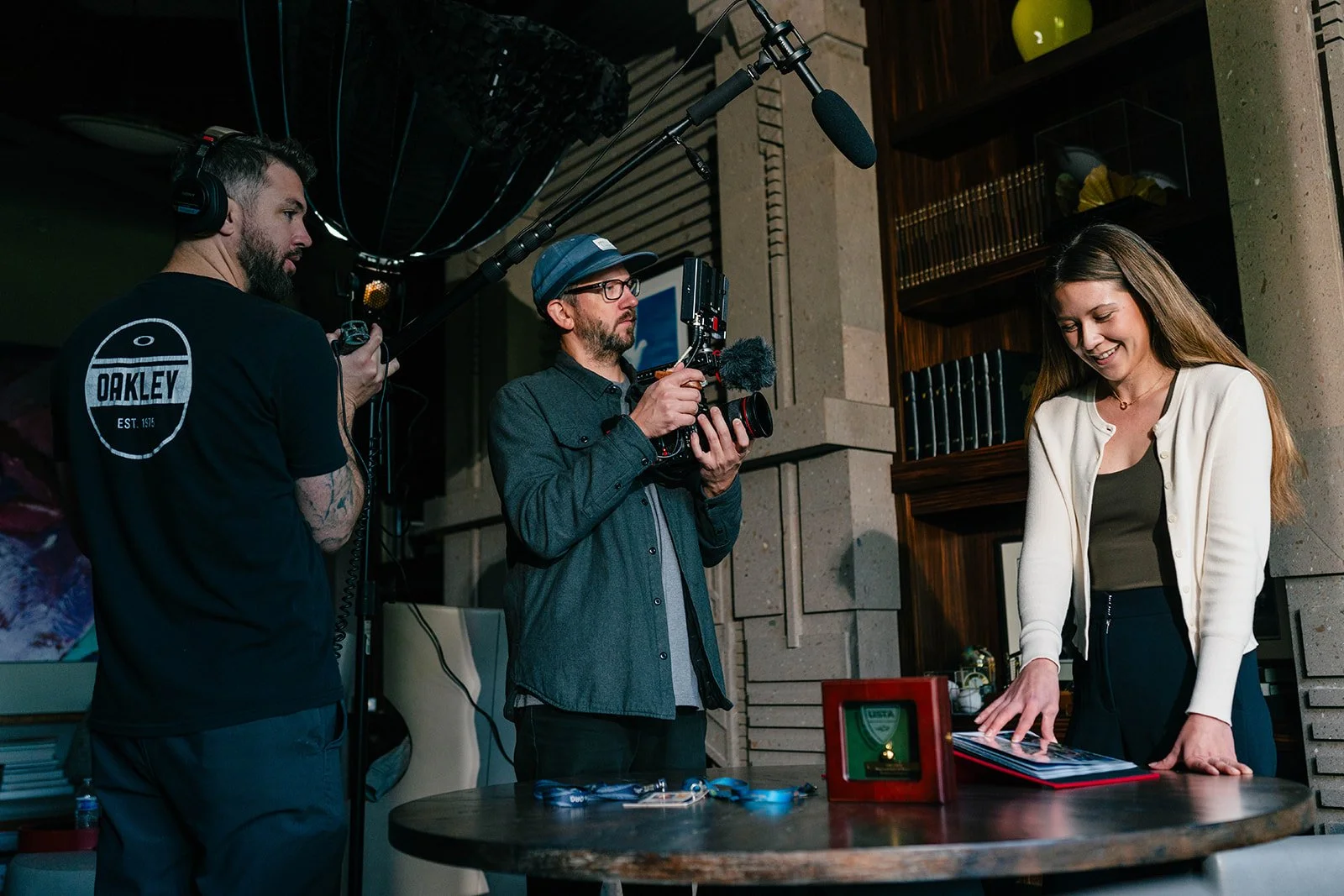 A young woman is smiling and looking down at a photo album or scrapbook on a table, while two men record her with a camera and microphone in a cozy, modern room with bookshelves and decorative items.