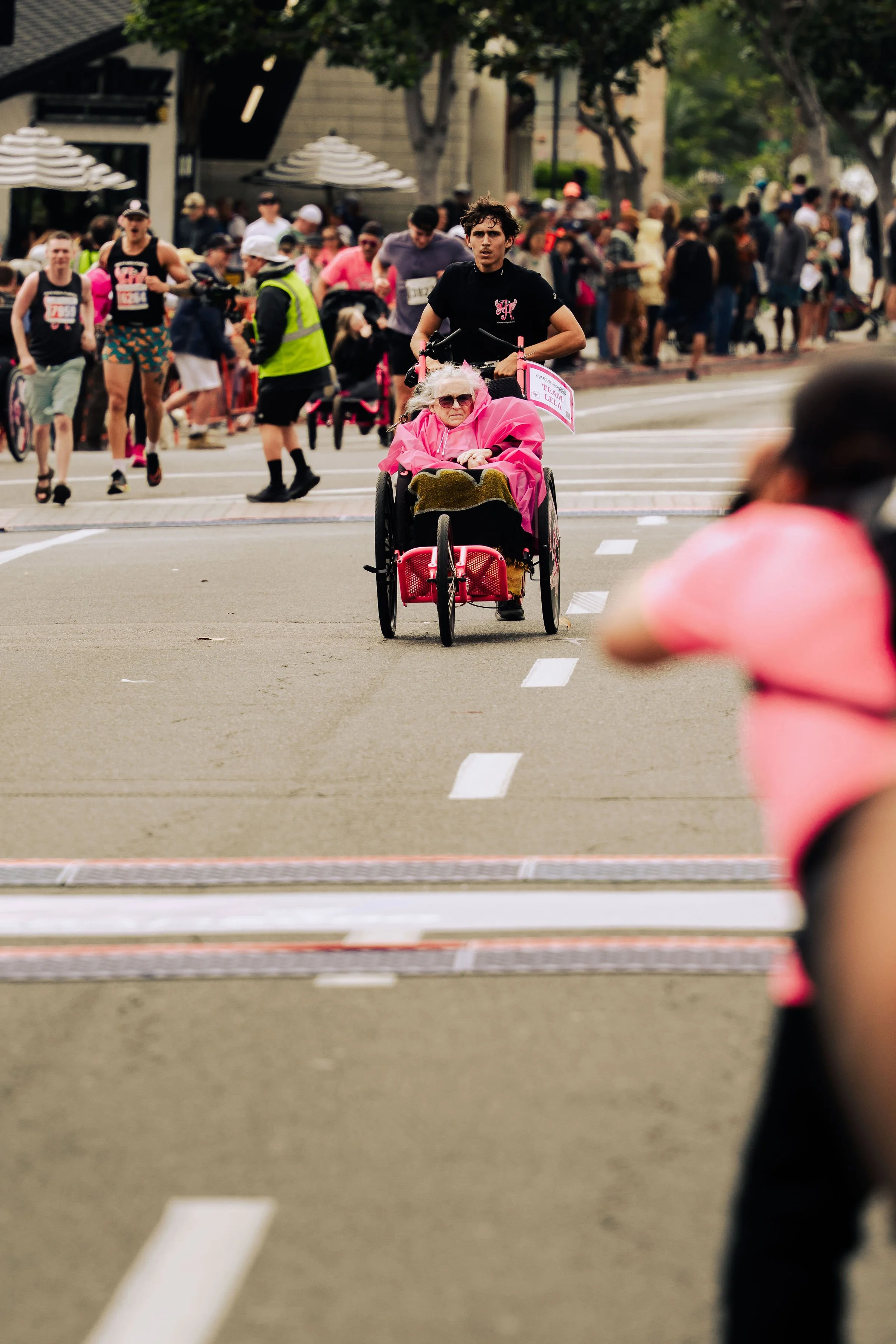 A person with gray hair and sunglasses sitting in a wheelchair wearing a bright pink jacket during a race or event, with a crowd of people in the background.