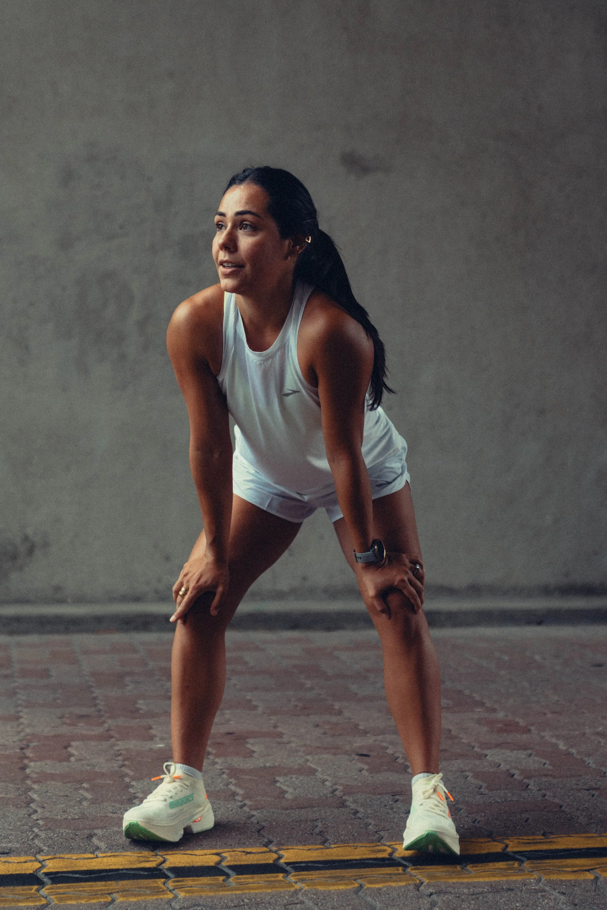 A woman in athletic clothing, wearing a white tank top, white shorts, and sneakers, is stretching outdoors on a paved surface with a gray wall in the background.
