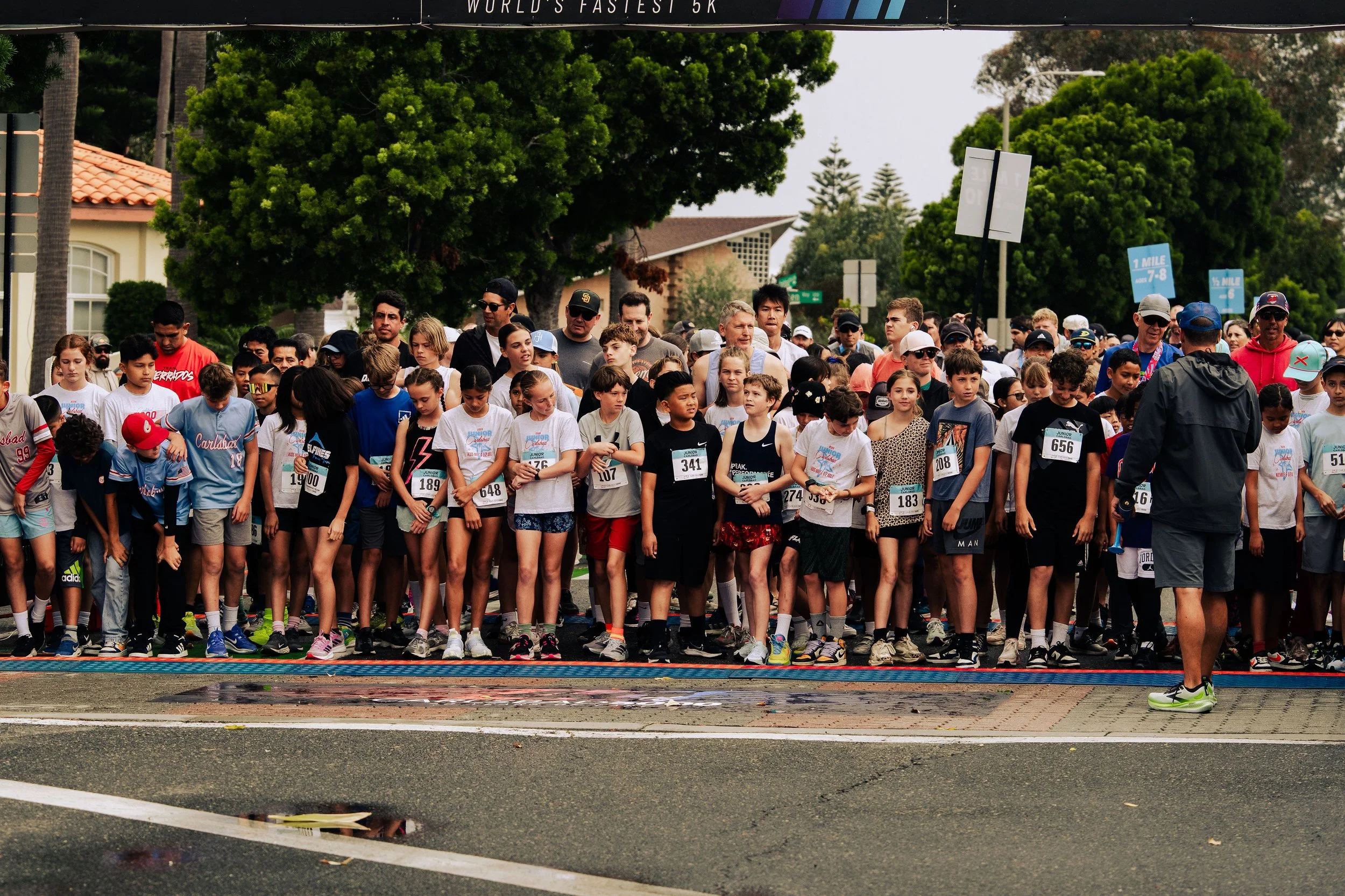 A large group of children and adults gathered at a starting line for a race, with some wearing race bibs and athletic clothing. Trees and residential houses are visible in the background.