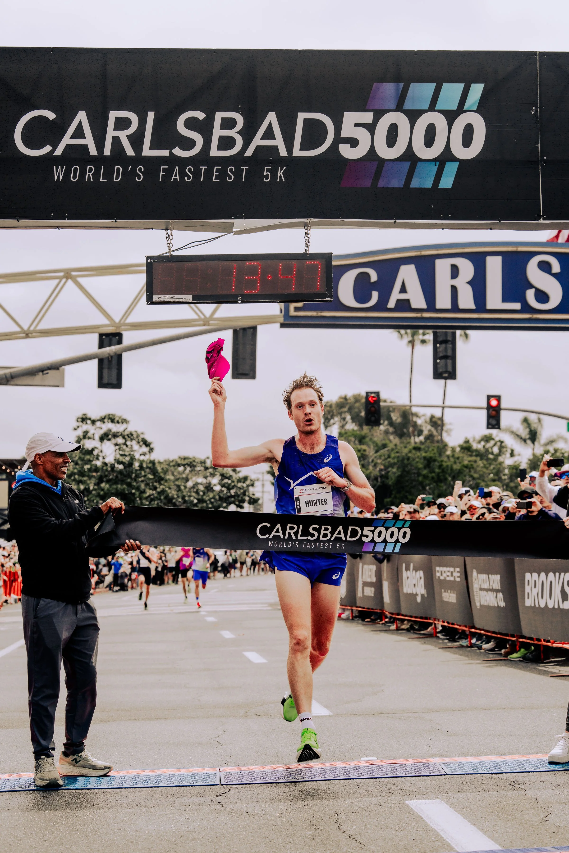 A male athlete in a blue running outfit and green shoes crosses the finish line of a race as a crowd cheers in the background. A large black banner above displays the race name 'CARLSBAD 5000' and 'WORLD'S FASTEST 5K'. The digital timer shows 13:47.