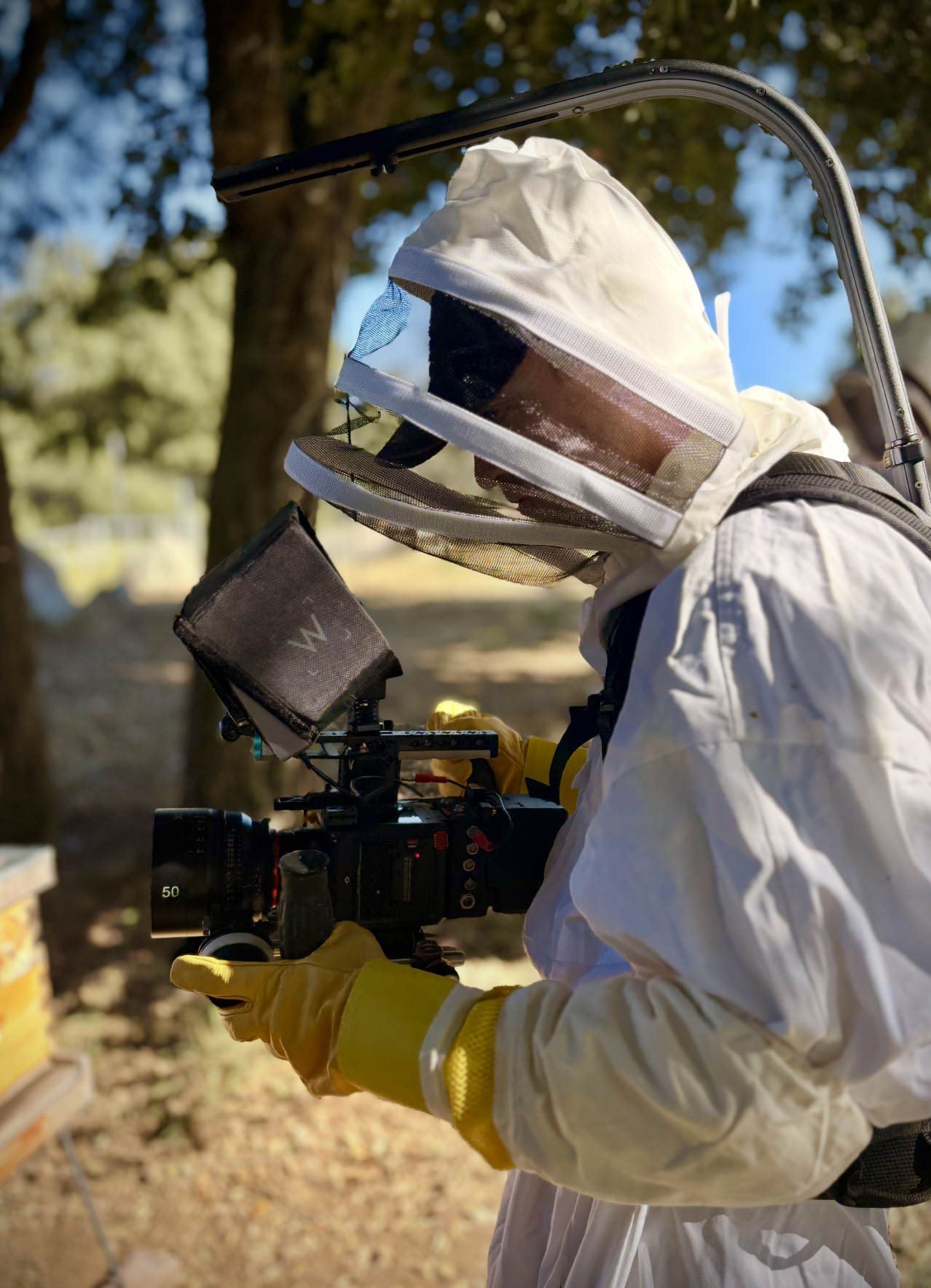 A scientist in a white protective suit with yellow gloves, wearing a protective face shield, working outdoors with a camera mounted on a tripod.