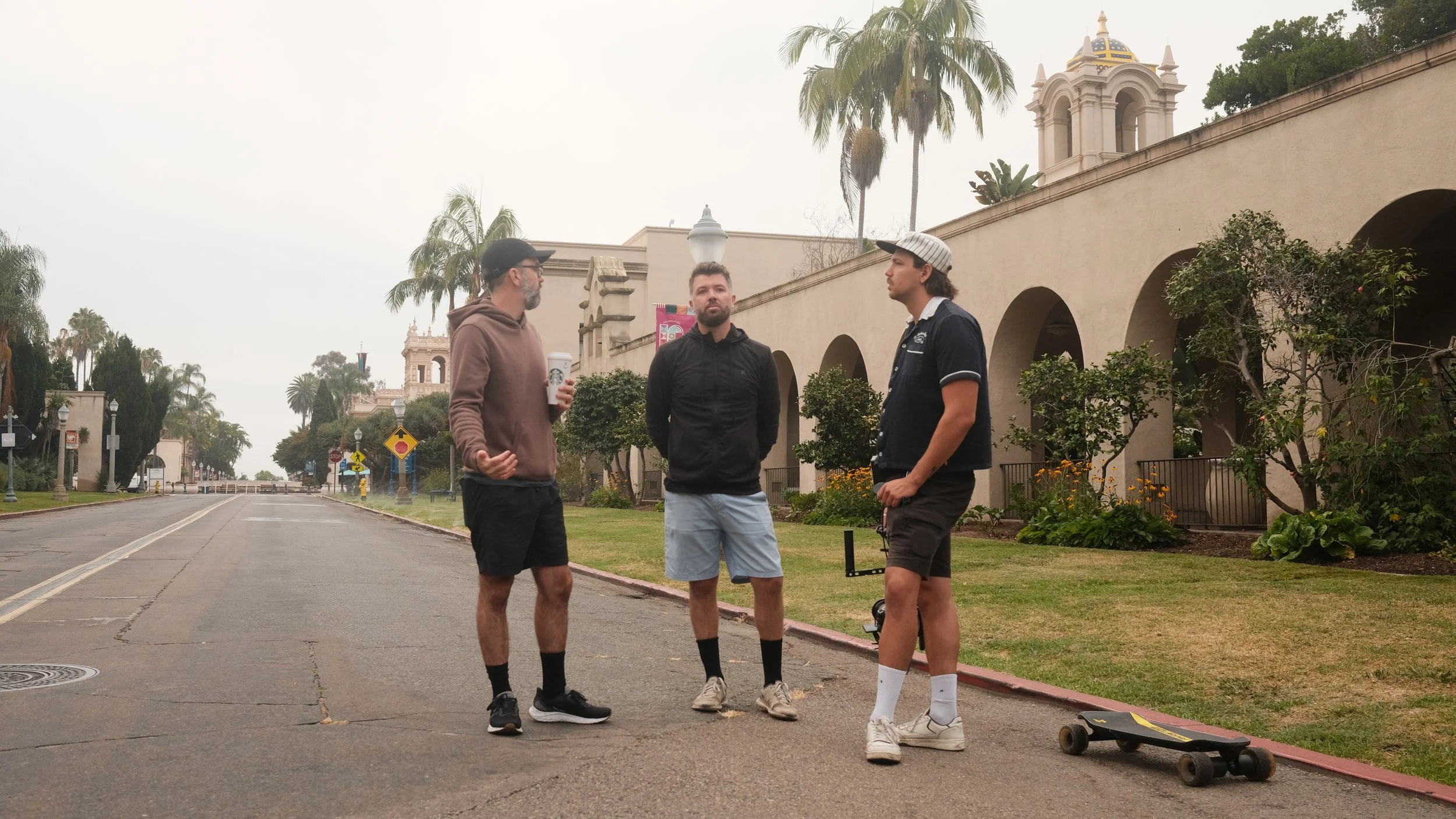 Three men stand on a sidewalk in a park near a building with arches and a tower, talking, with a skateboard on the ground nearby.