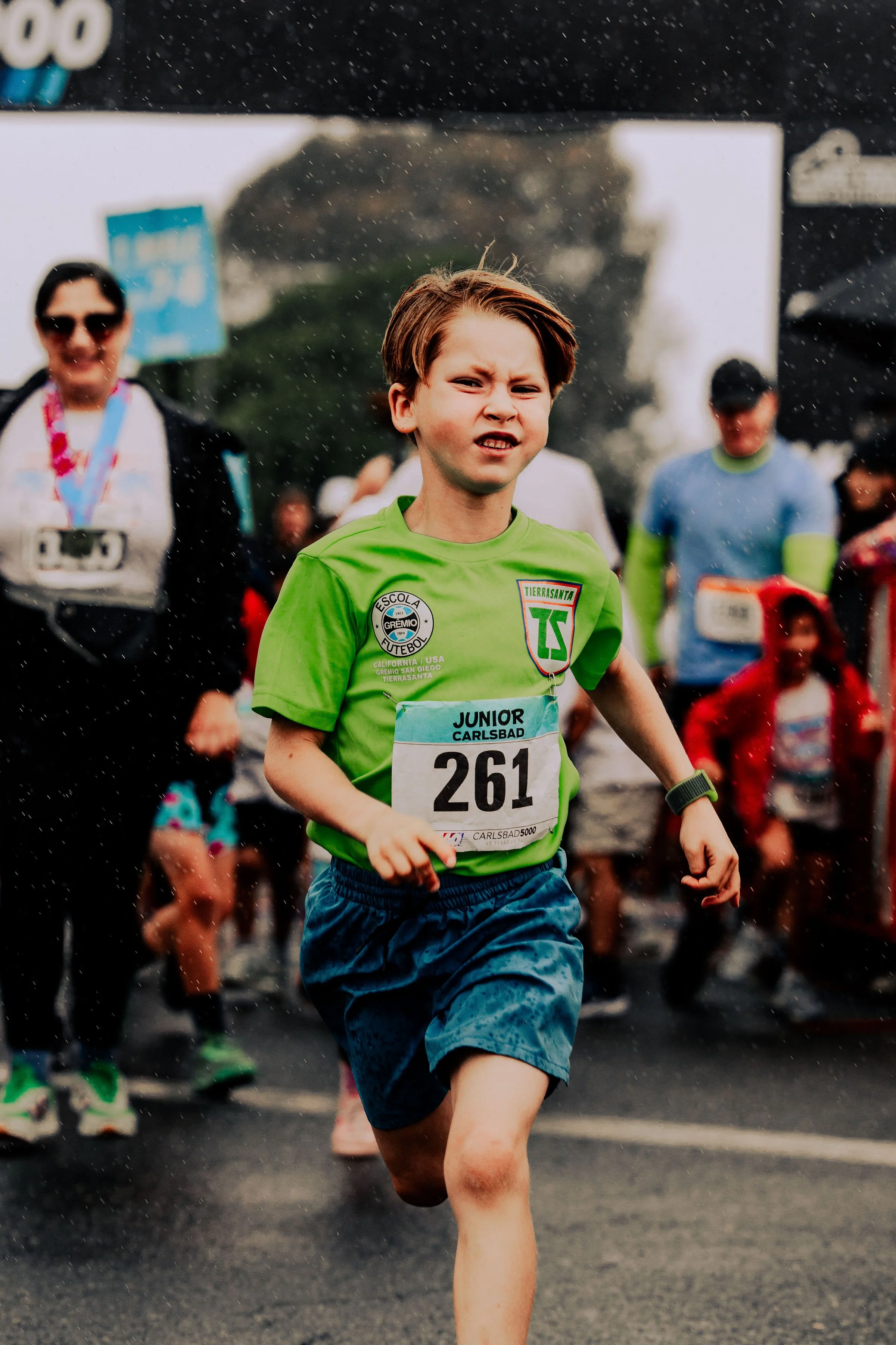 A young boy running in a race wearing a green shirt with a bib number, with people running behind him. It appears to be a race event in the rain.