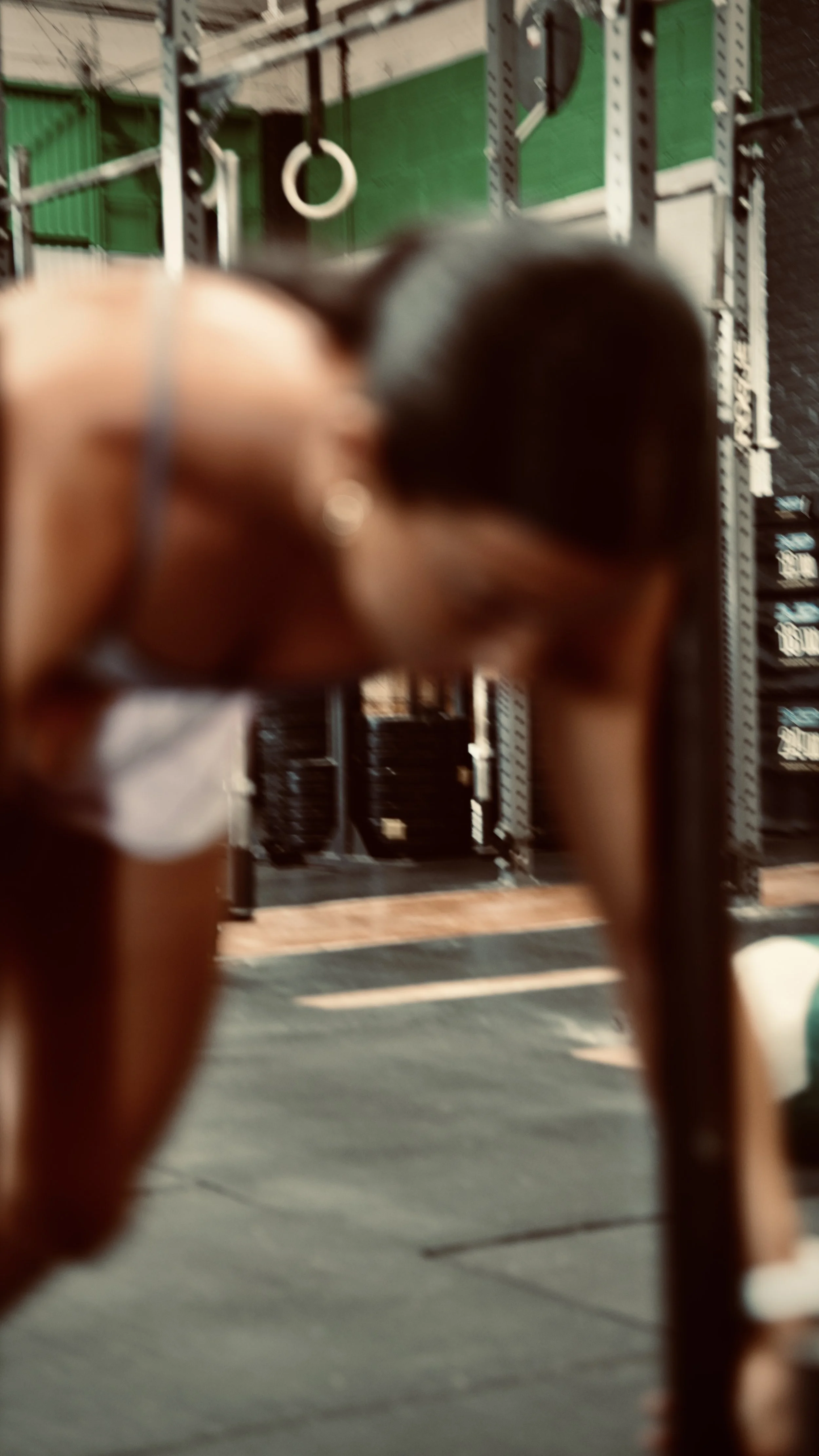 A person is in a gym, bending forward with their back to the camera. The background features gym equipment such as pull-up rings, a green wall, and metal gym racks.