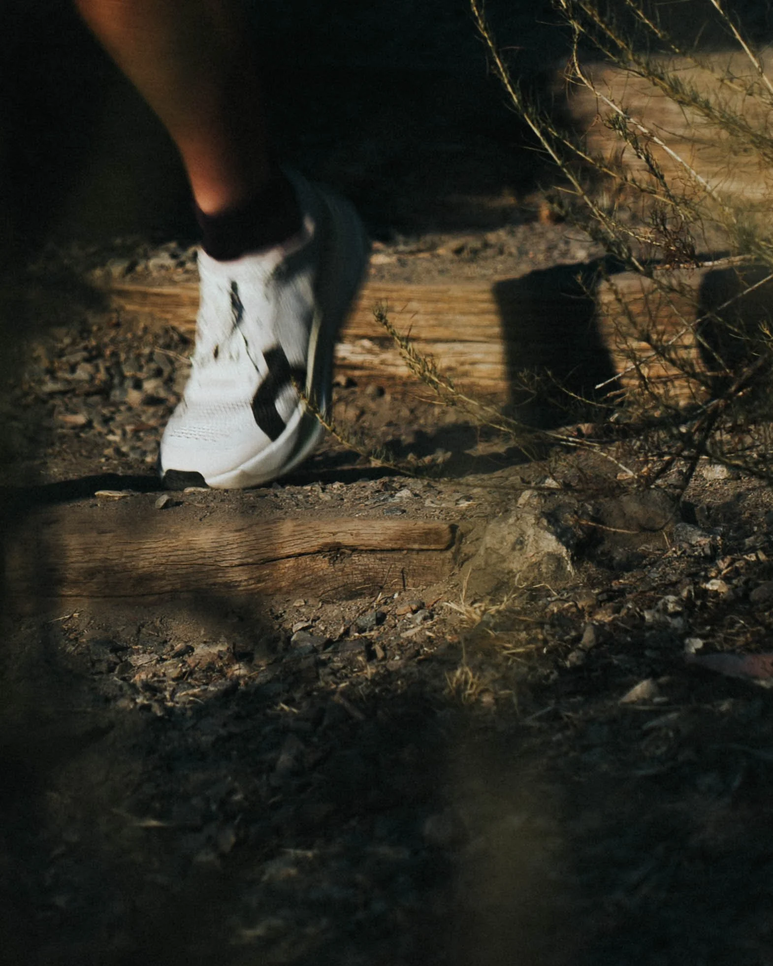 Close-up of a person tying their white sneaker on a rugged, dirt outdoor trail at night, with dry grass and small rocks visible around.