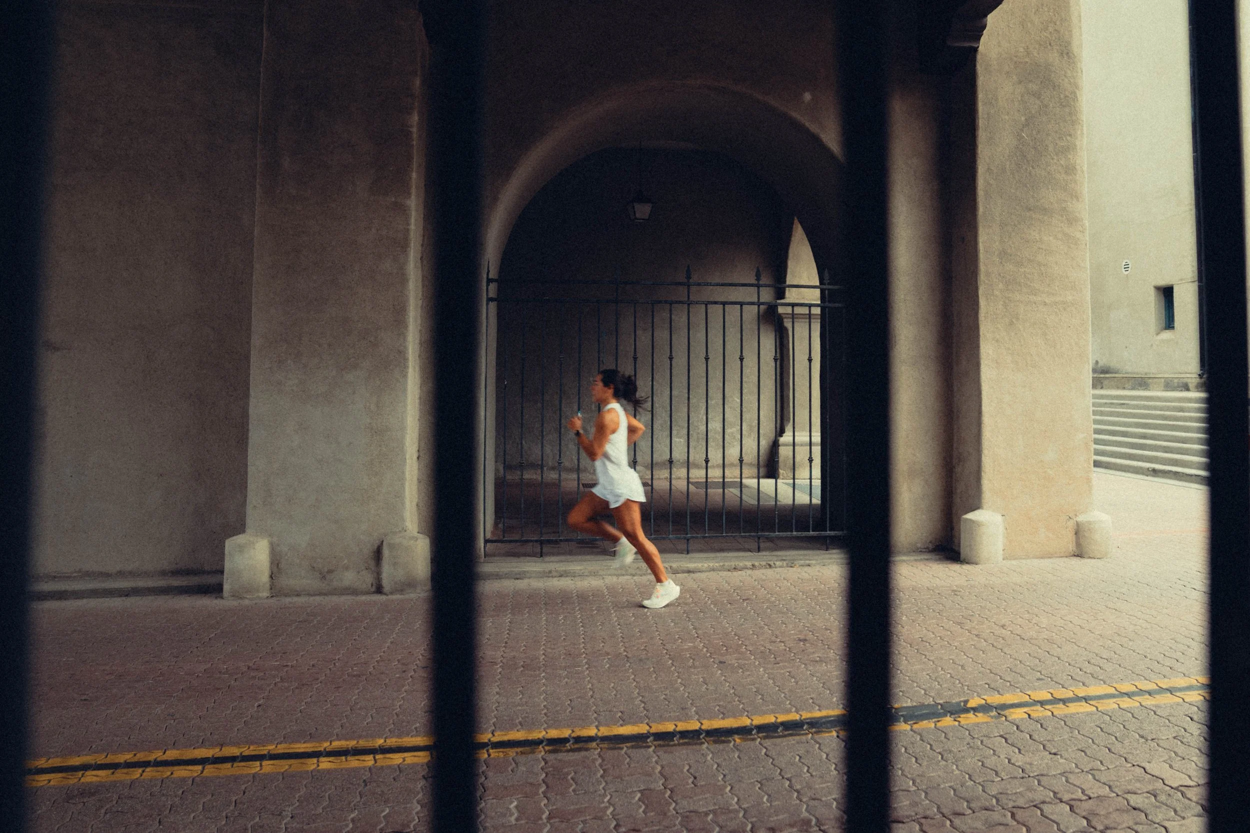 A woman jogging or running past a gated archway in an urban setting, viewed through black bars in the foreground.