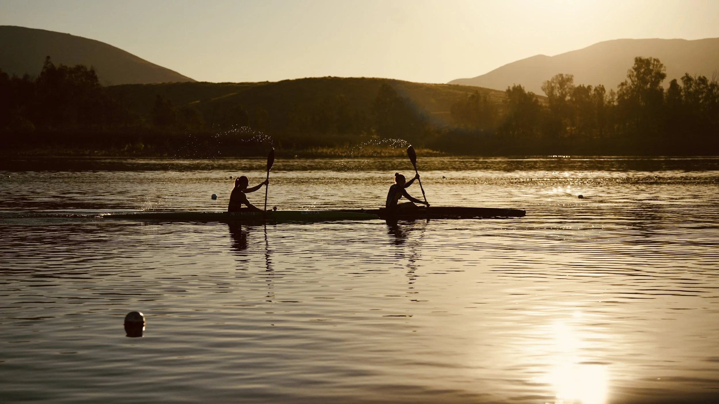 Two people kayaking on a lake during sunset with mountainous landscape in the background.