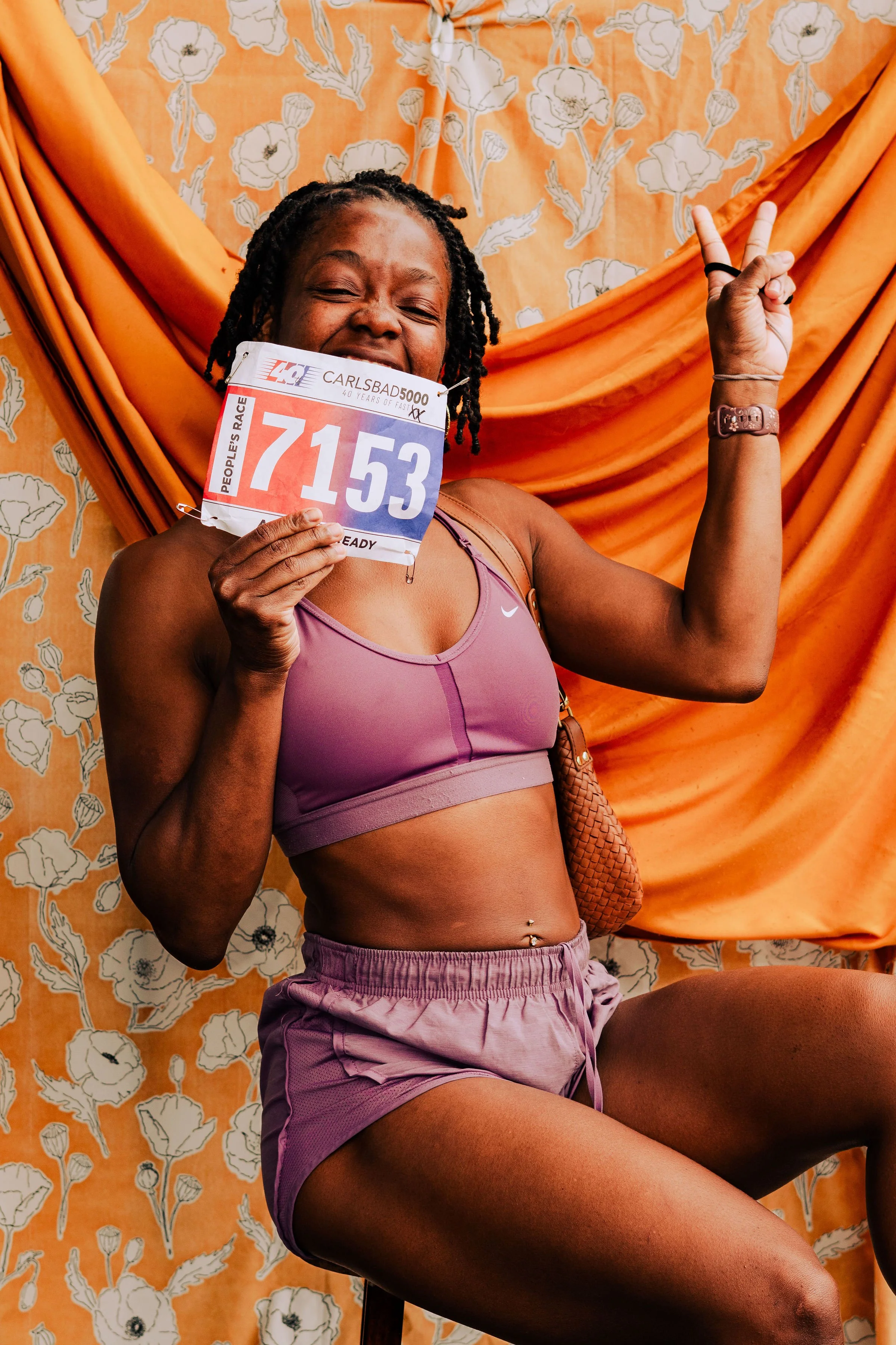 A woman wearing a purple sports bra and shorts holds a race bib and makes a peace sign with her right hand, sitting against an orange floral backdrop.