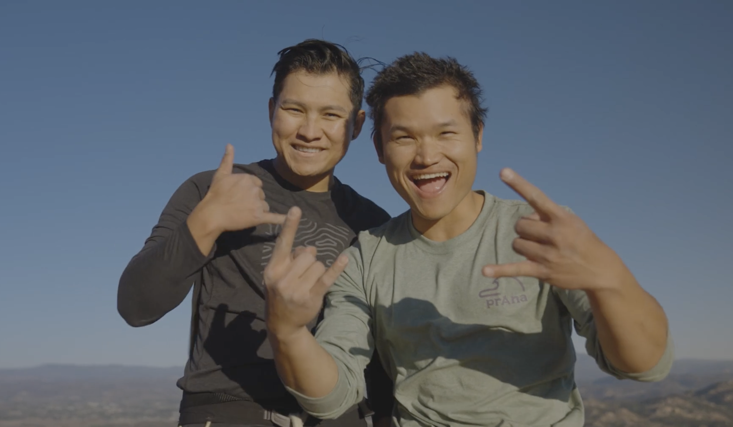 Two smiling young men outdoors against a clear blue sky, making hand signs with their fingers, one with a dark long-sleeve shirt and the other with a light T-shirt.