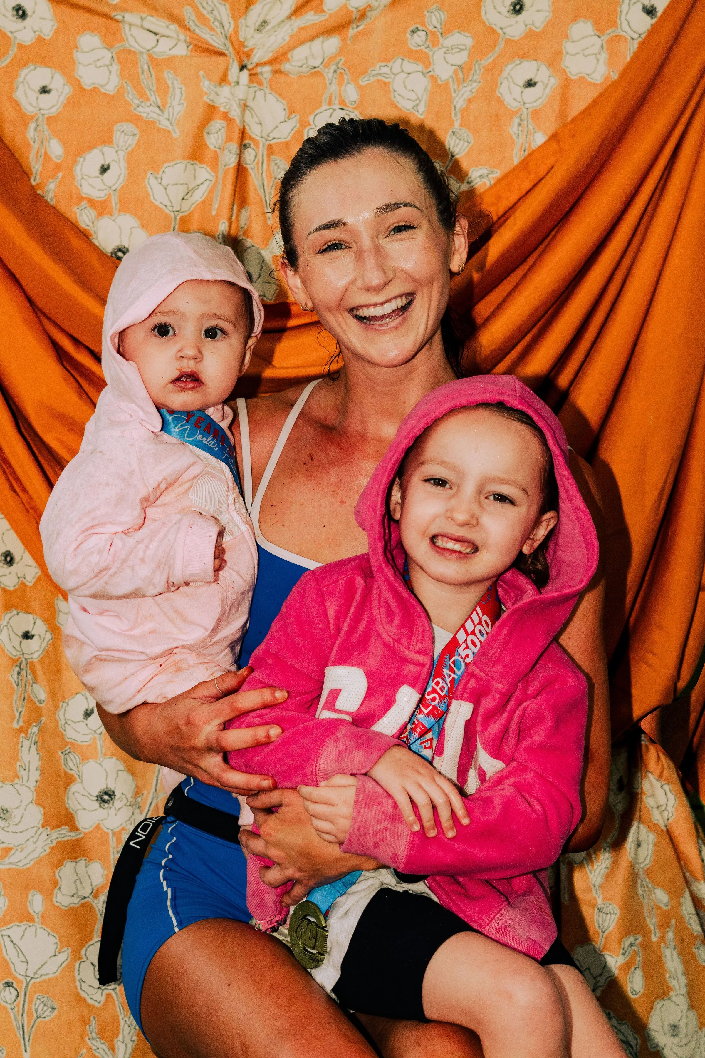 A woman smiling and holding two young children, one in each arm, against an orange floral backdrop.