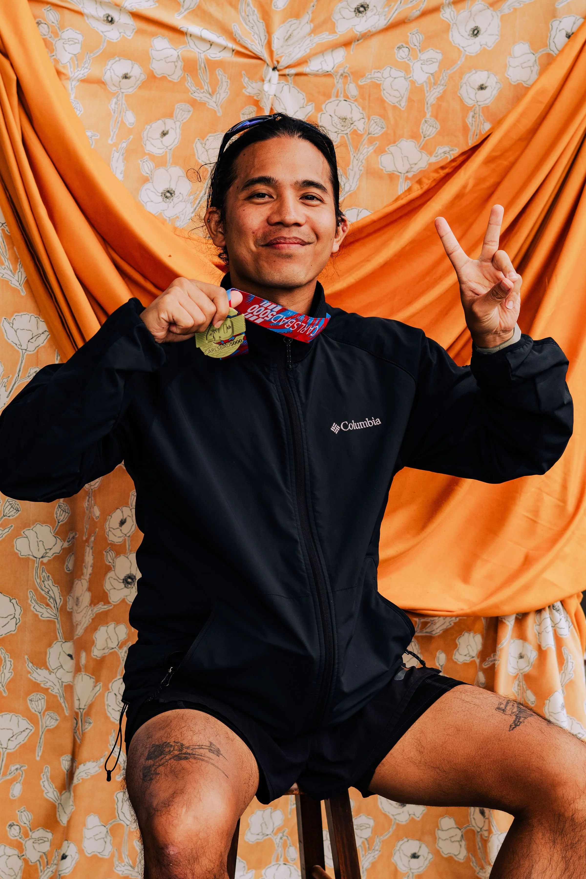 A man sitting on a stool in front of orange floral curtains, holding a marathon medal and making a peace sign with his right hand, wearing a black Columbia jacket and black shorts.