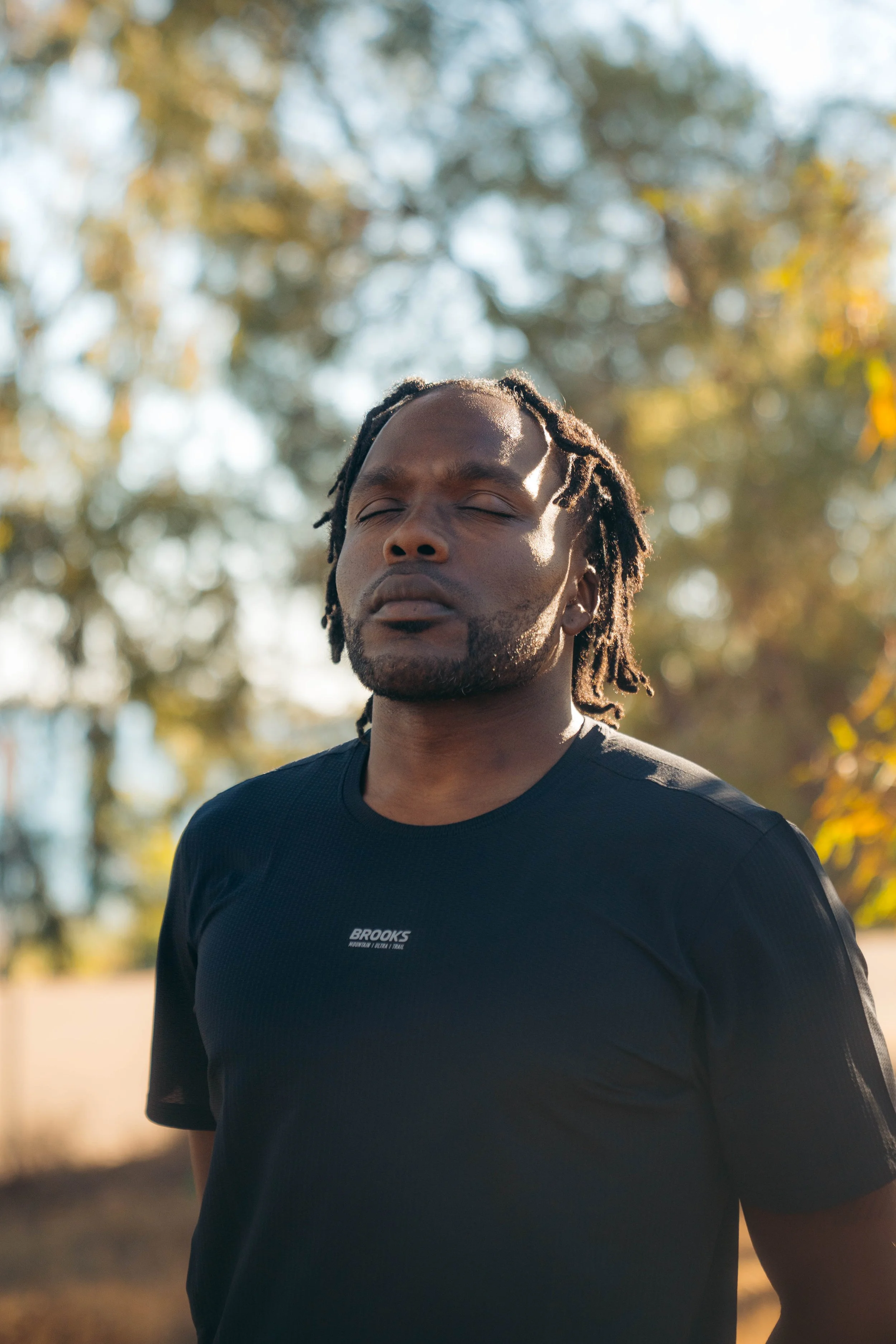 A man with closed eyes standing outdoors in a park with sunlight filtering through trees, wearing a black Brooks athletic shirt.