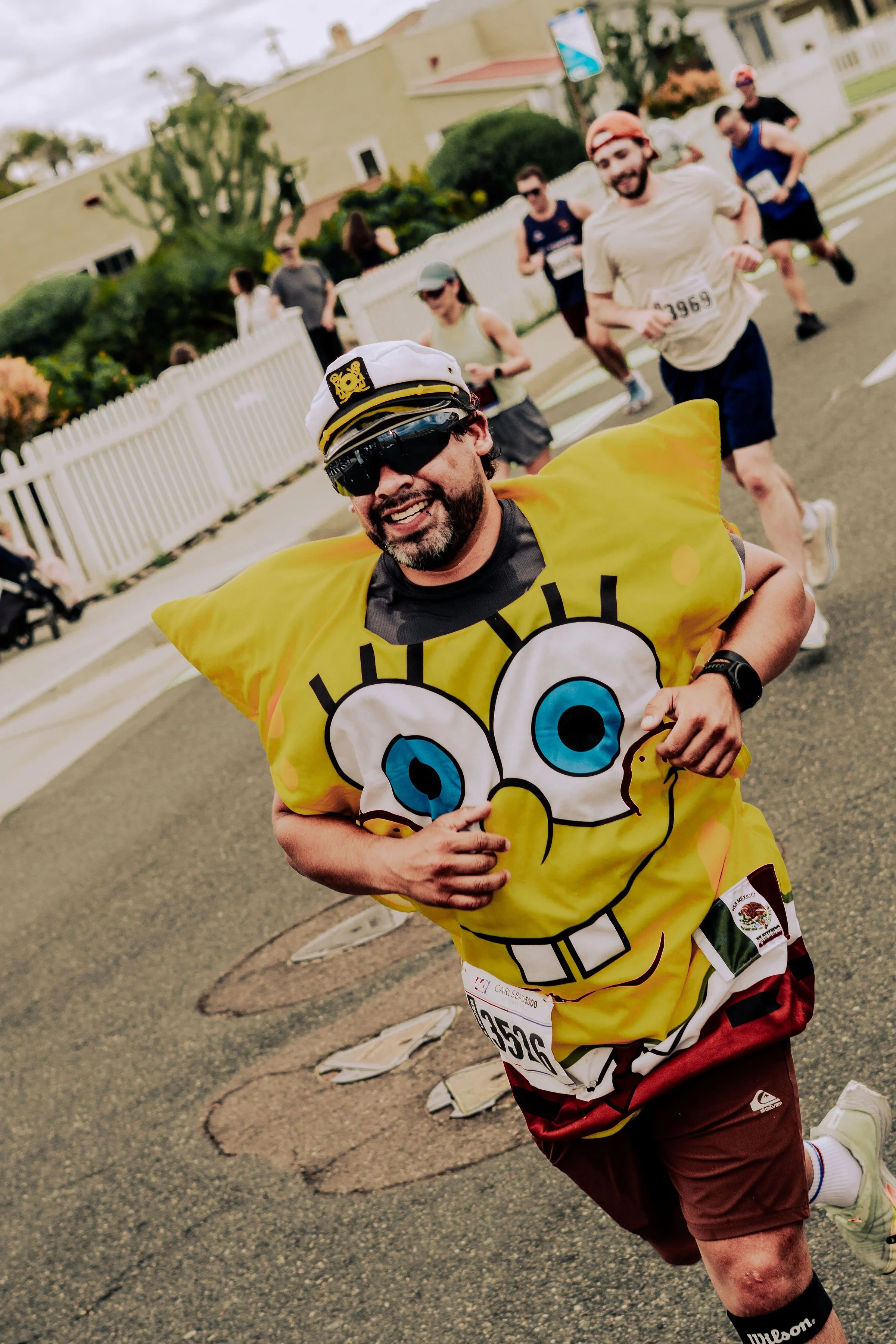 A group of runners participating in a race on a street with white fence, some houses, and greenery in the background. The runner in front is dressed as SpongeBob SquarePants.