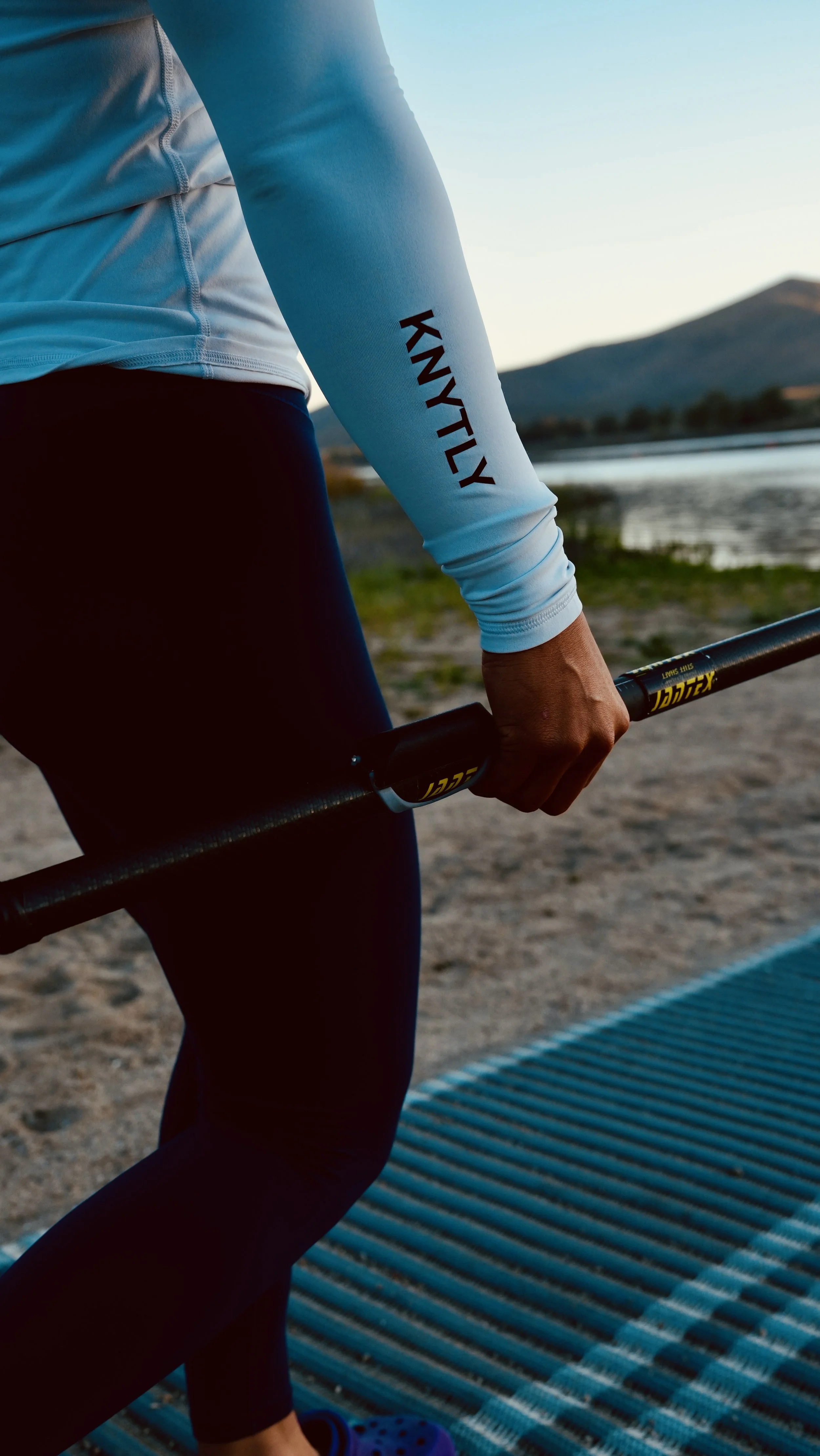 Person holding a black barbell with yellow writing on the handle, wearing a white long-sleeve shirt with 'KNYTLY' on the sleeve and black leggings, outdoors near water and mountains at sunset.