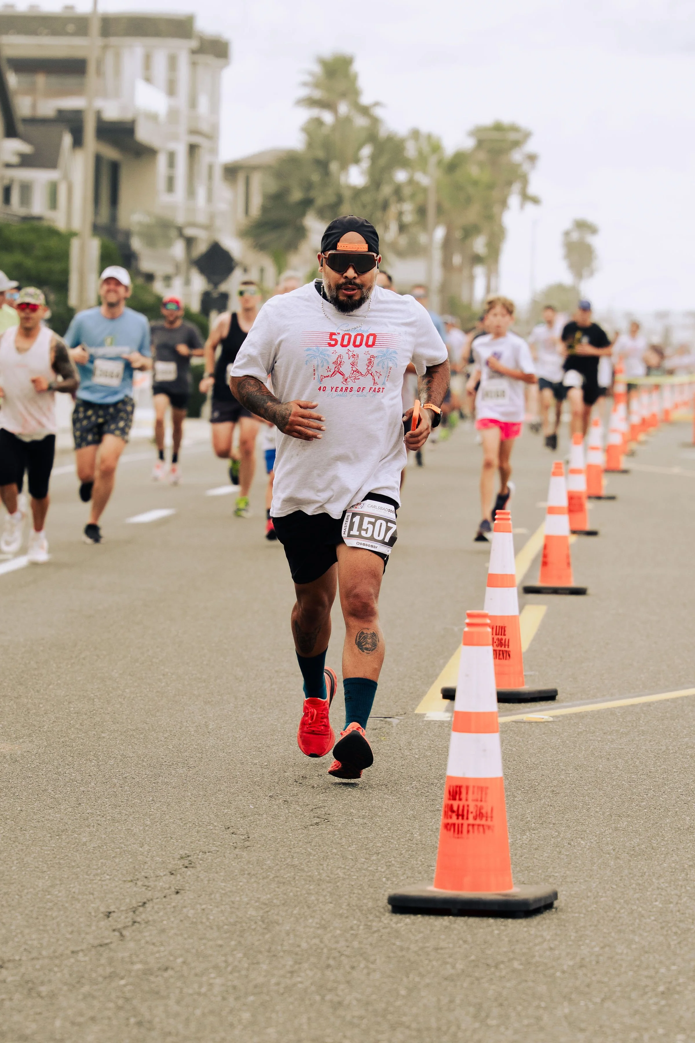 A man running in a race, wearing a white T-shirt, black shorts, red running shoes, sunglasses, and a black cap. Several other runners are visible in the background, along with orange traffic cones marking the race course alongside a road, with reside