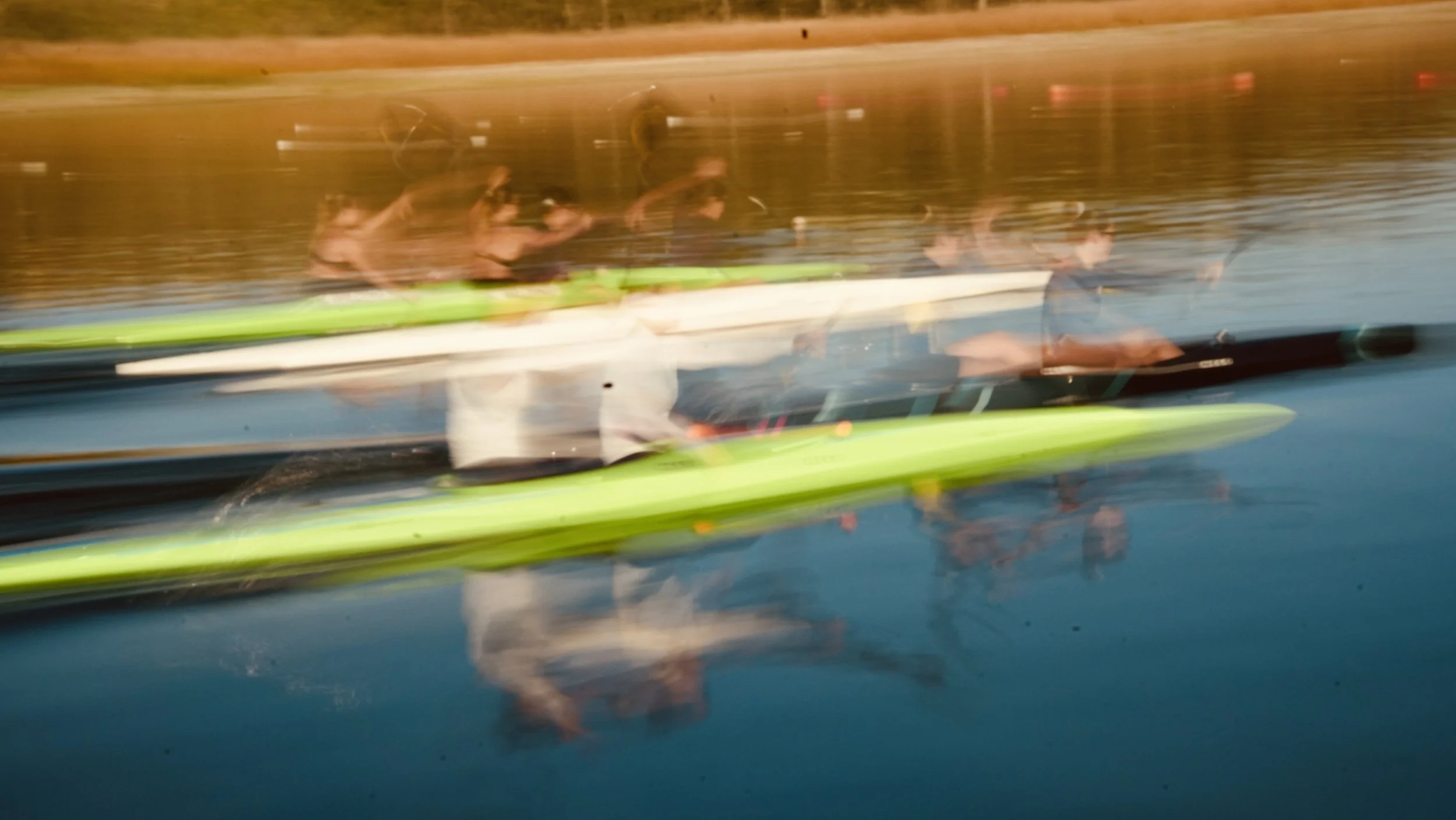 A blurred photo of three people in a boat or kayak on a body of water, with a background of a shoreline with orange and brown tones.