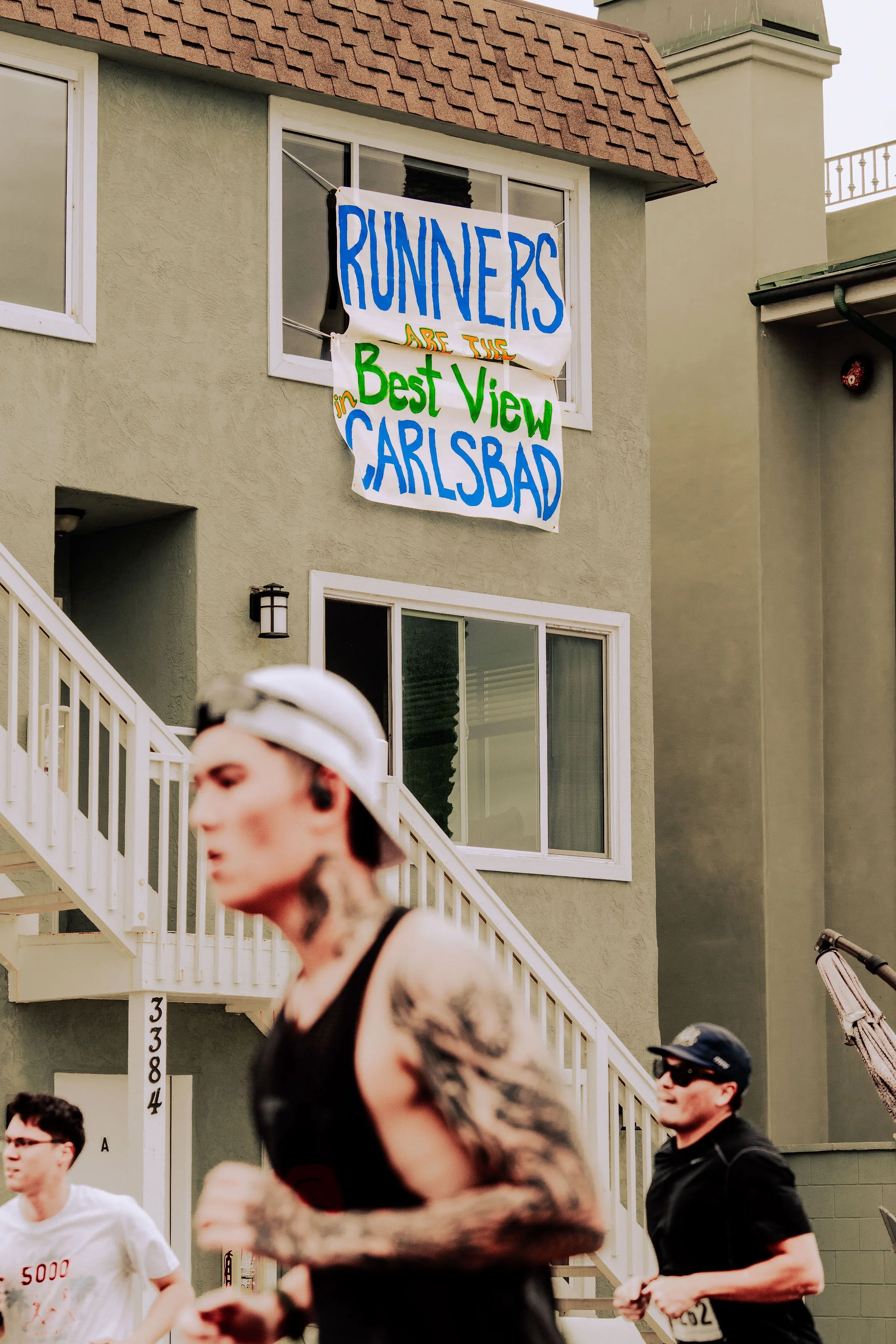 A group of runners participating in a race, with a building in the background displaying a sign that reads 'RUNNERS ARE THE BEST VIEW IN CARLSBAD.'