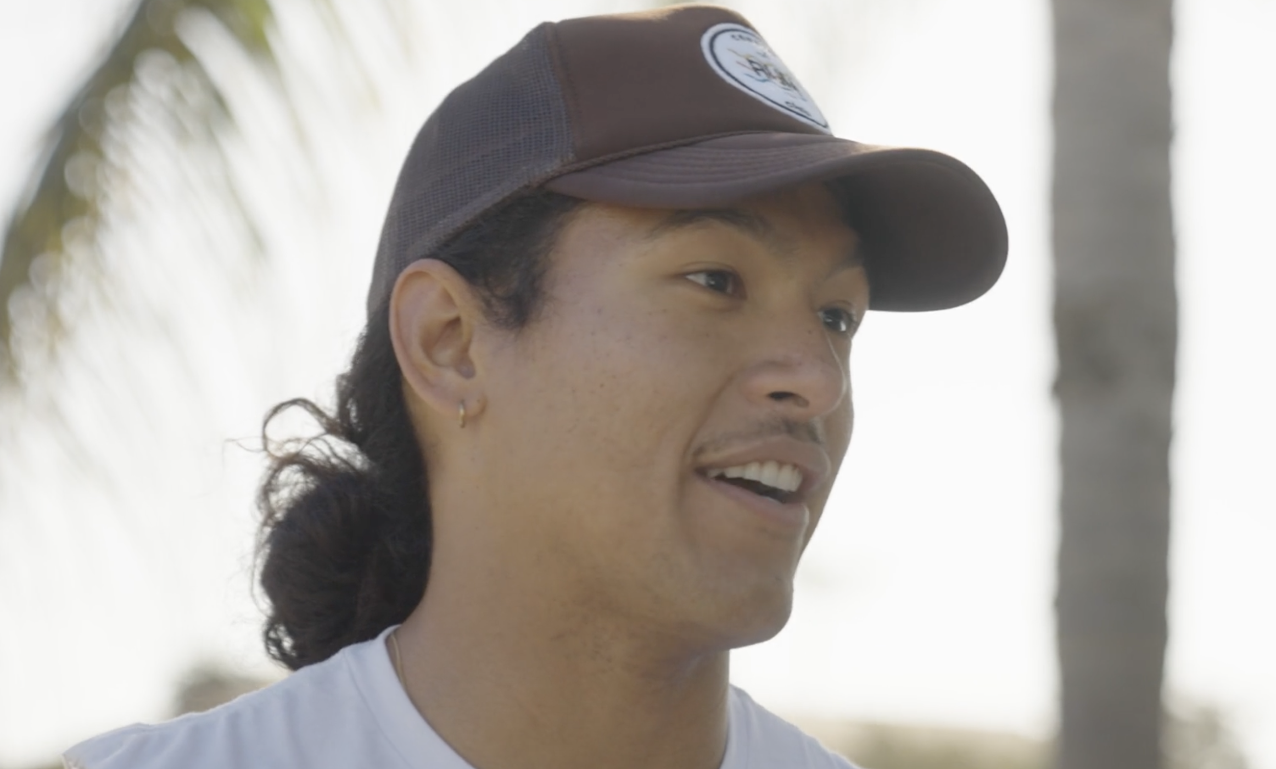 Close-up of a smiling young man with long dark hair, wearing a brown cap and a white shirt, outdoors near palm trees.