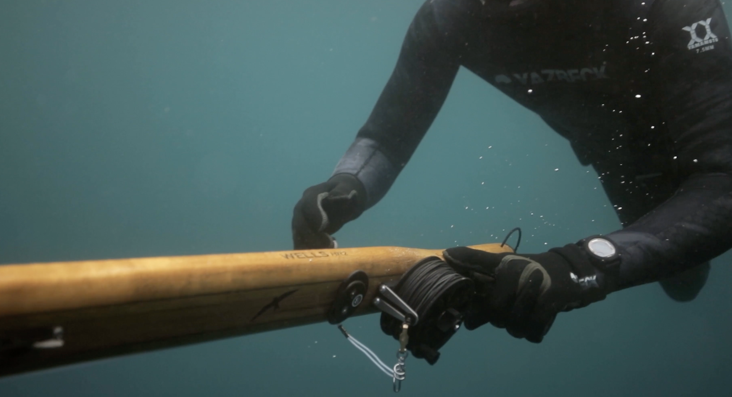 Person wearing a wetsuit and gloves holding a wooden paddle underwater.